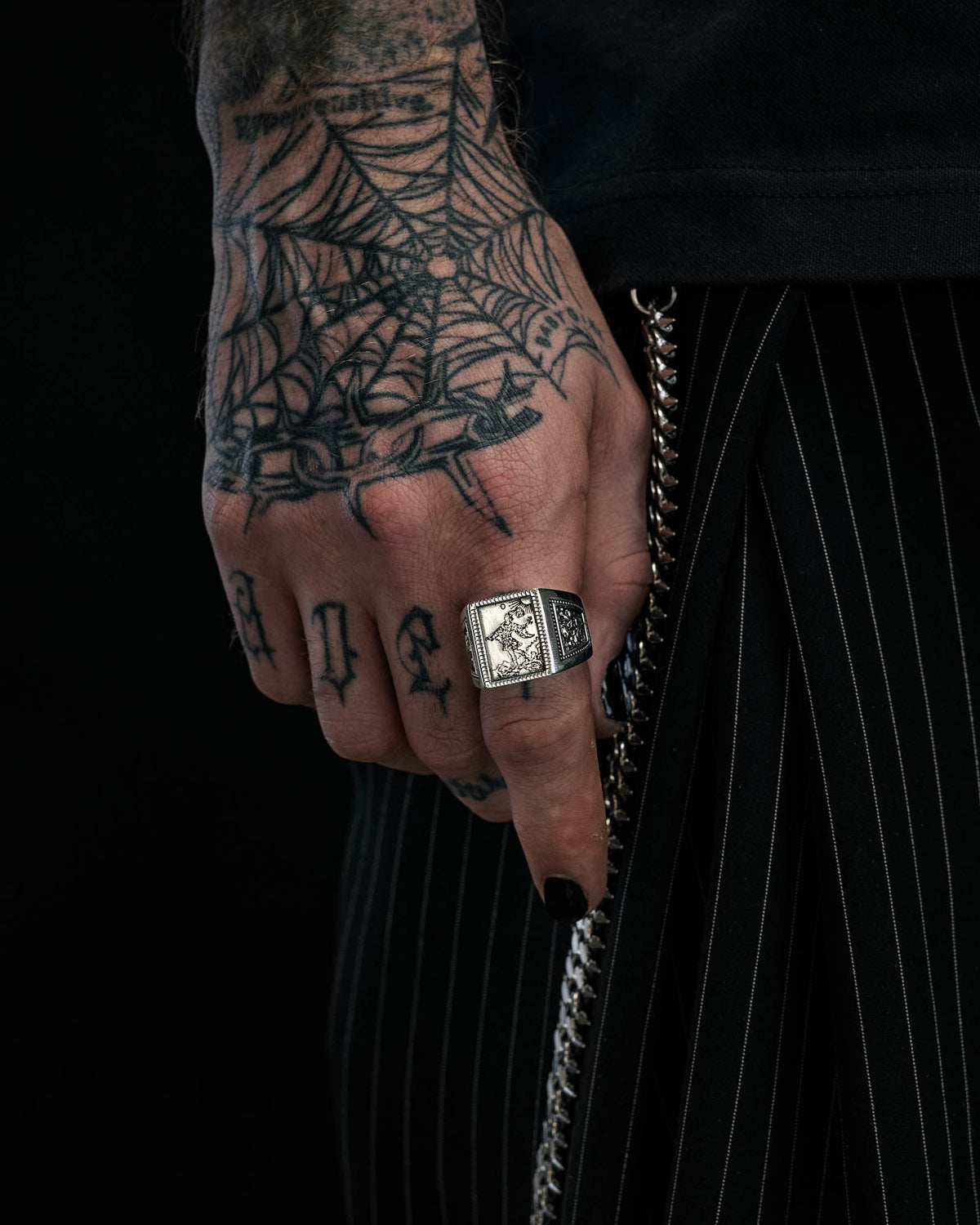 A tattooed hand with a spiderweb tattoo, wearing the Statement Collective 925 Sterling Silver “Try Hard Fool” Ring and black nail polish, rests against pinstriped pants with a visible chain.