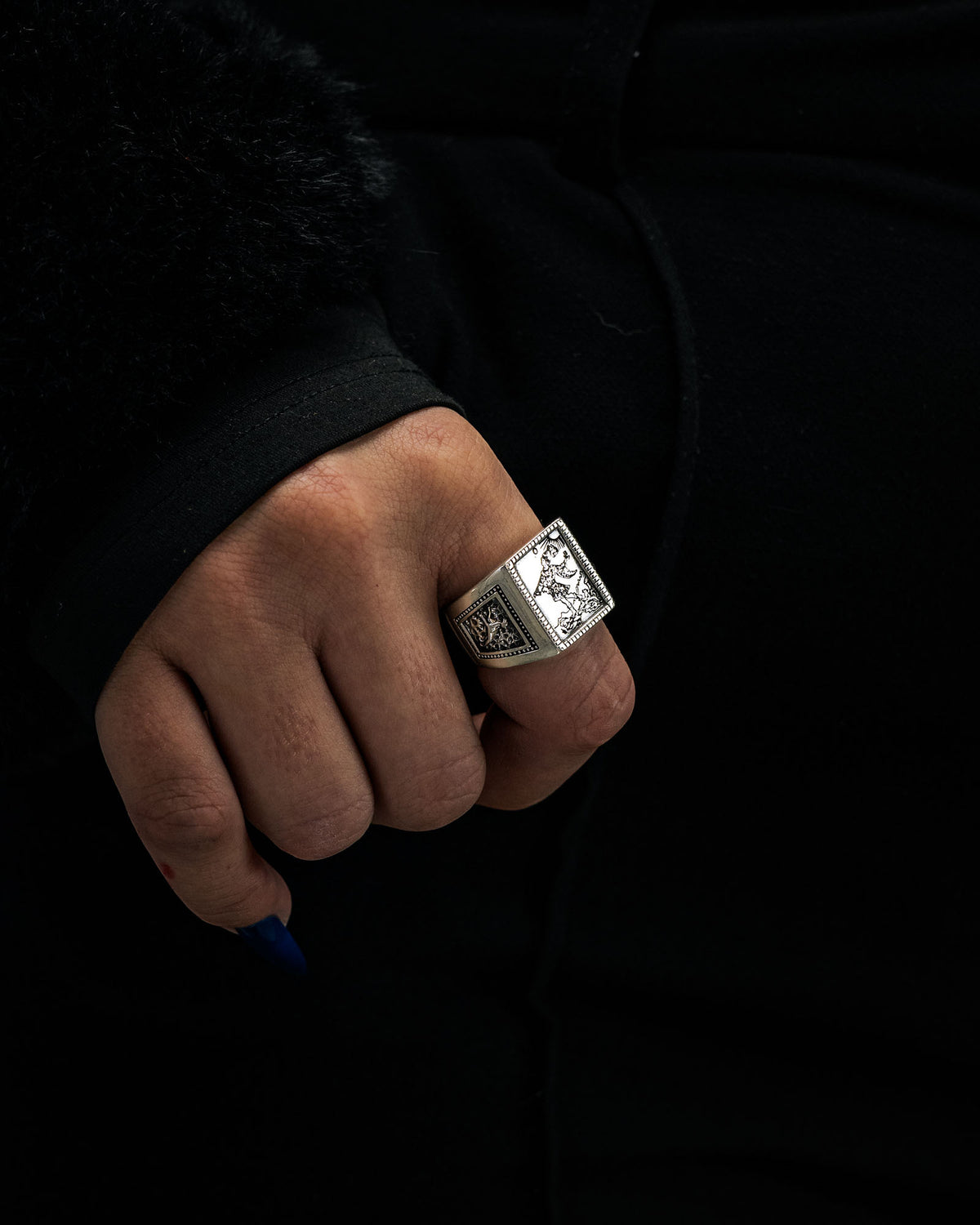 A hand wearing the Statement Collective 925 Sterling Silver “Try Hard Fool” Ring, featuring a bold square design and intricate engraving, is shown against dark clothing and a shadowy background.