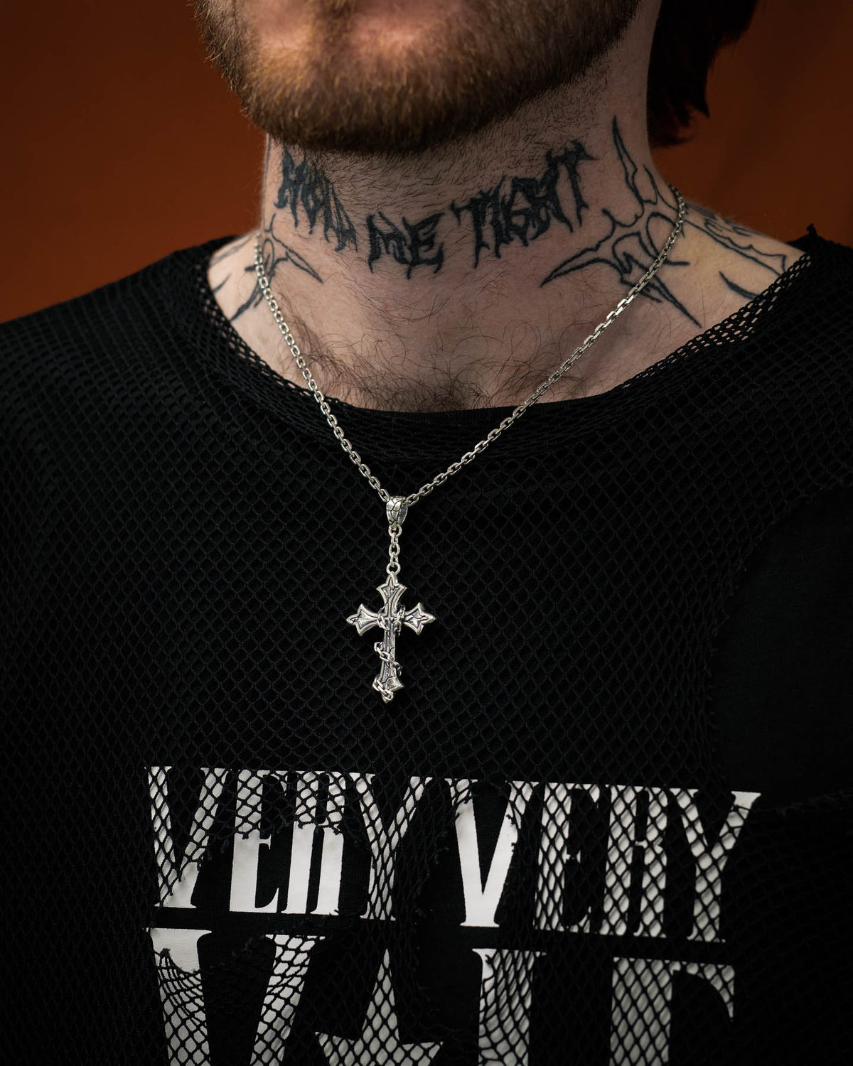 A person with a neck tattoo wears Statement Collective’s 925 Sterling Silver Mini “The Ruins” Cross Pendant and a black mesh shirt featuring bold white text that reads VERY VERY, blending modern edge with striking gothic jewelry style.