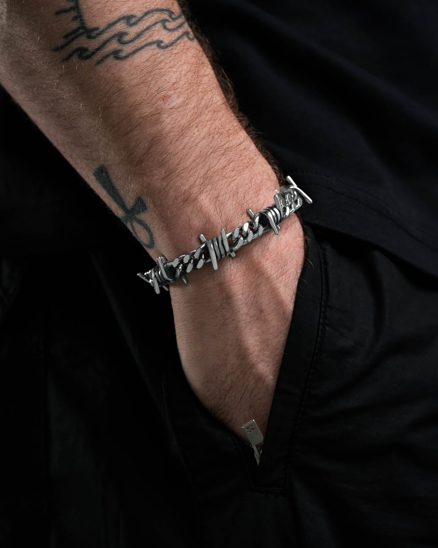 A close-up shows a person’s hand in their pocket, wearing the edgy 9mm Barbed Wire Cuban Bracelet. Their arm displays an ankh and wavy line tattoo, while black clothing highlights the bold barbed wire jewelry style.