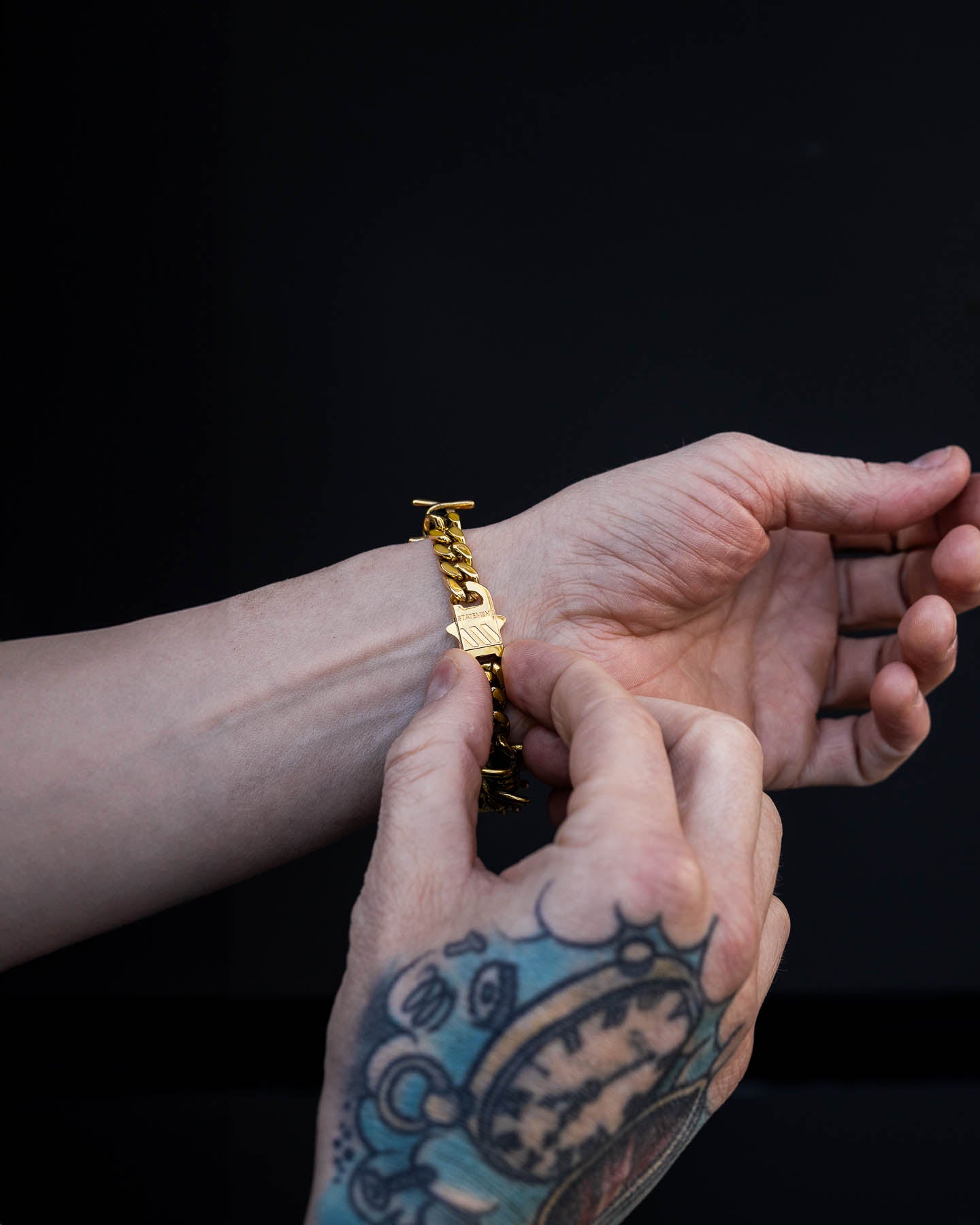A person with a tattooed hand fastens the 9mm Barbed Wire Cuban Bracelet (Gold) on their other wrist against a plain black background.