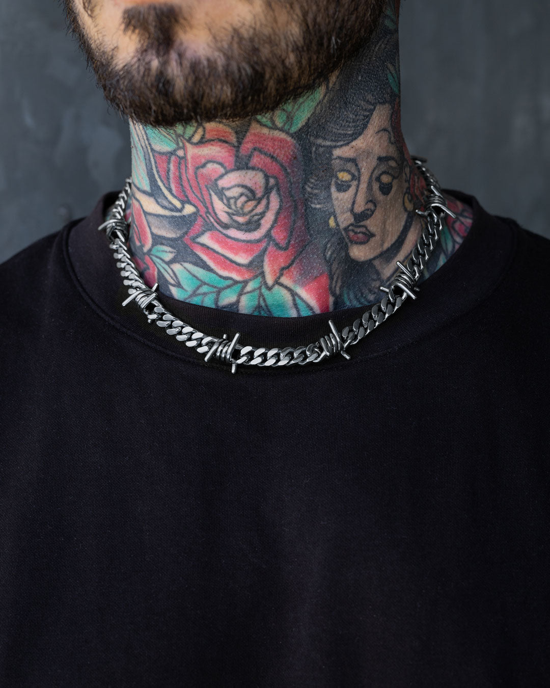 A person wearing a black shirt, the 9mm Barbed Wire Cuban Chain necklace, and colorful neck tattoos with a large rose and a woman’s face.