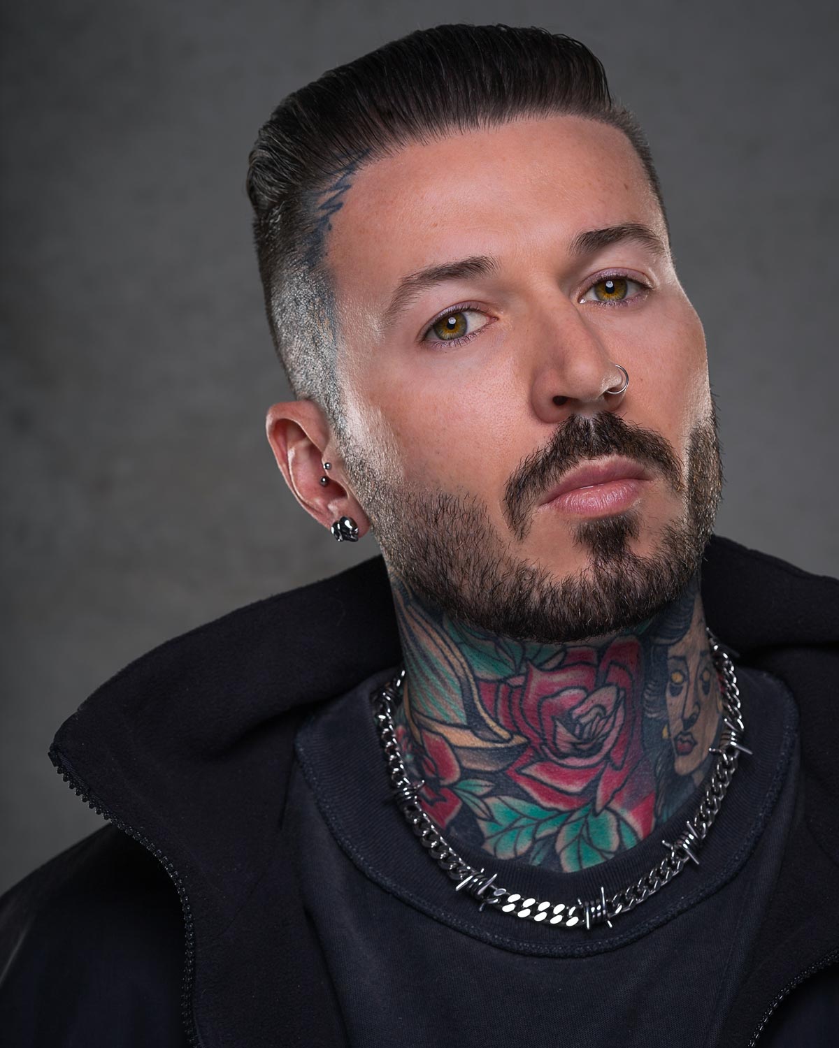 A man with slicked-back hair, trimmed beard, and nose piercing looks at the camera. He has neck tattoos—roses and a woman's face—and wears bold earrings, a dark jacket, and a 9mm Barbed Wire Cuban Chain.