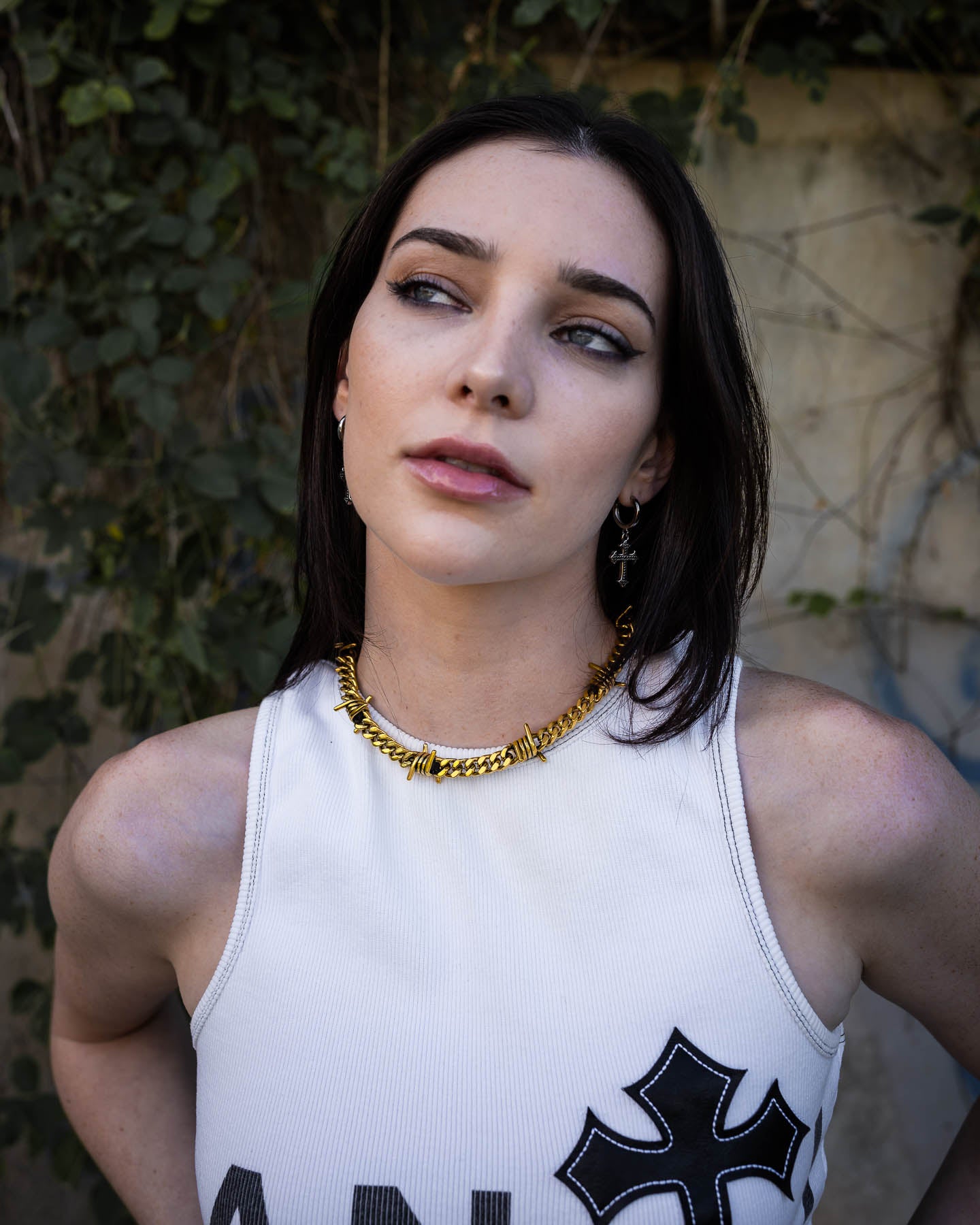 A woman with straight dark hair, wearing a white sleeveless top with a black cross patch and the 9mm Barbed Wire Cuban Chain (Gold), stands outdoors before green ivy, gazing upward with a calm expression.