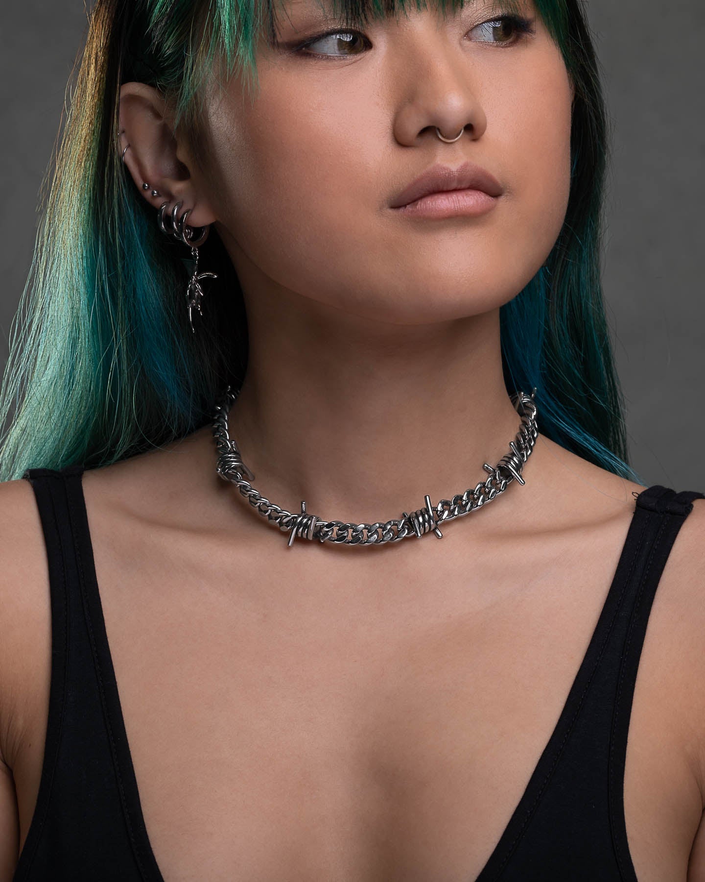 A person with teal and black hair wears the 9mm Barbed Wire Cuban Chain, silver earrings, a black tank top, and has multiple ear piercings plus a septum ring. They look slightly to the side against a gray background.