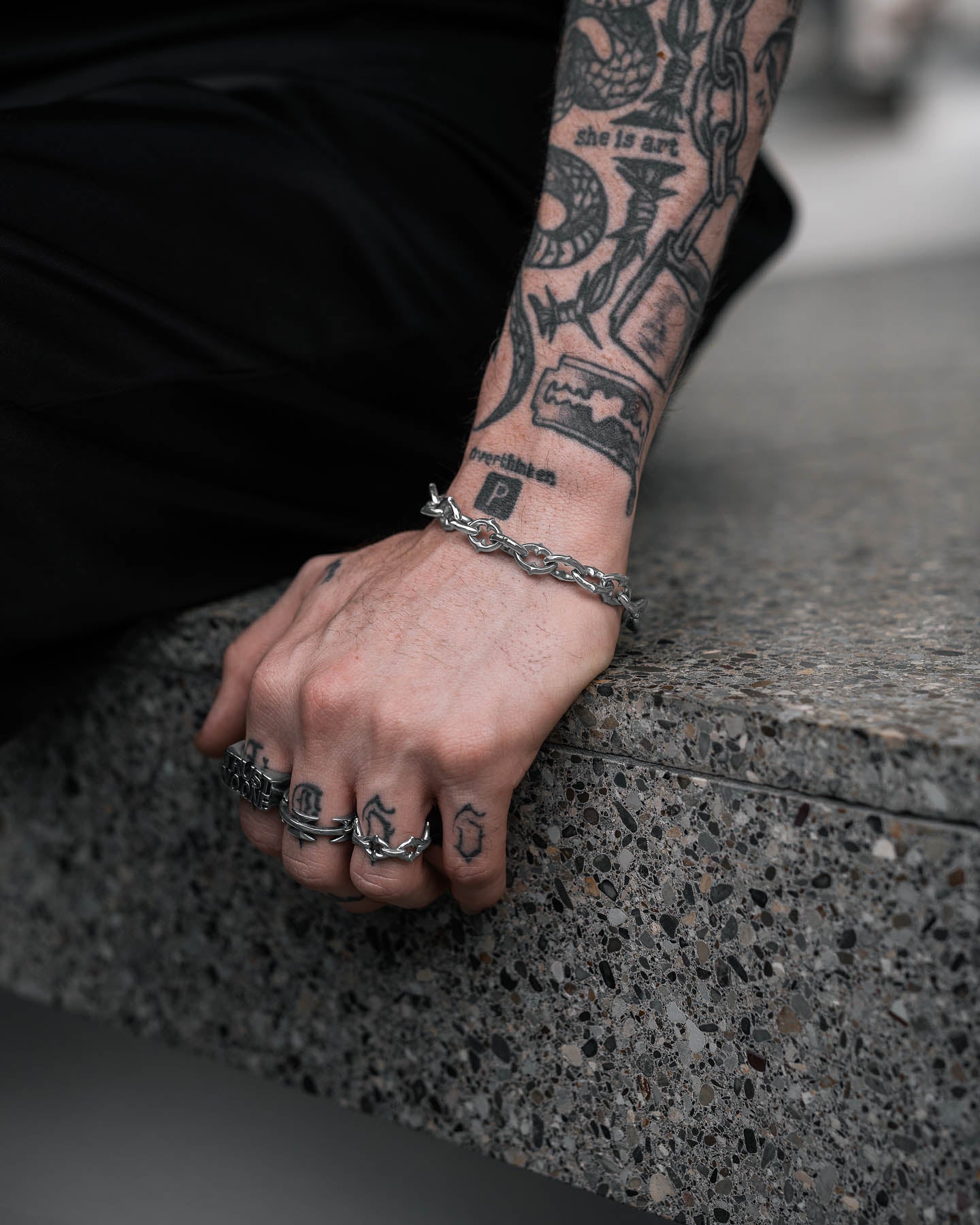 A close-up of a tattooed hand wearing rings and "The Cathedral" 9mm Spiked Bracelet, a non-tarnish, waterproof chain with medieval spikes, elegantly displayed on stone.