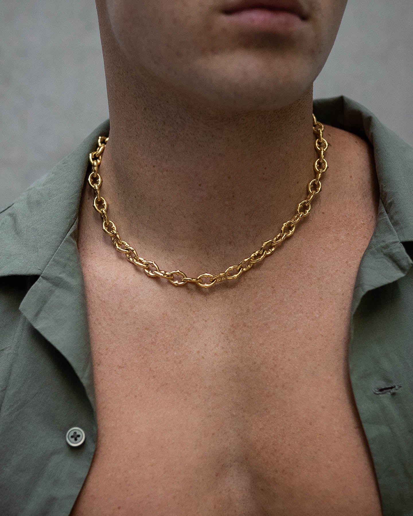 A person with a bare chest wears "The Cathedral" 9mm Spiked Chain (Gold) necklace over an open green collared shirt. The background is neutral and out of focus.