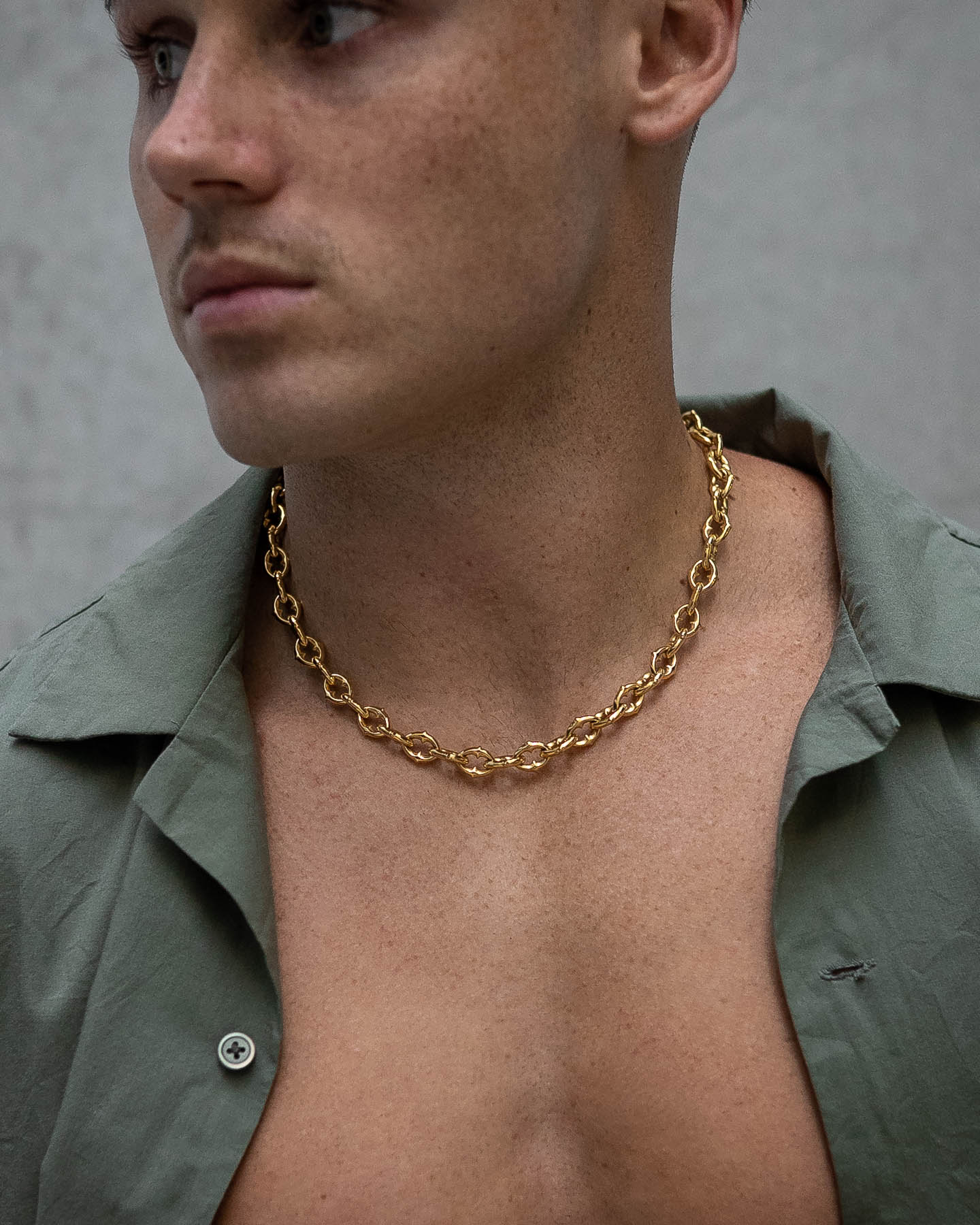 A person with short hair wears "The Cathedral" 9mm Spiked Chain (Gold) necklace and an open green shirt, exposing their chest, while looking off to the side against a plain background.