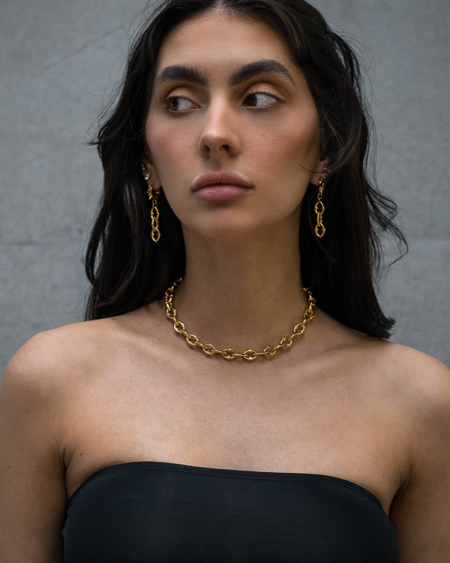 A woman with long dark hair wears a strapless black top and "The Cathedral" 9mm Spiked Chain (Gold) necklace and earrings, standing against a plain gray background while looking to her left.