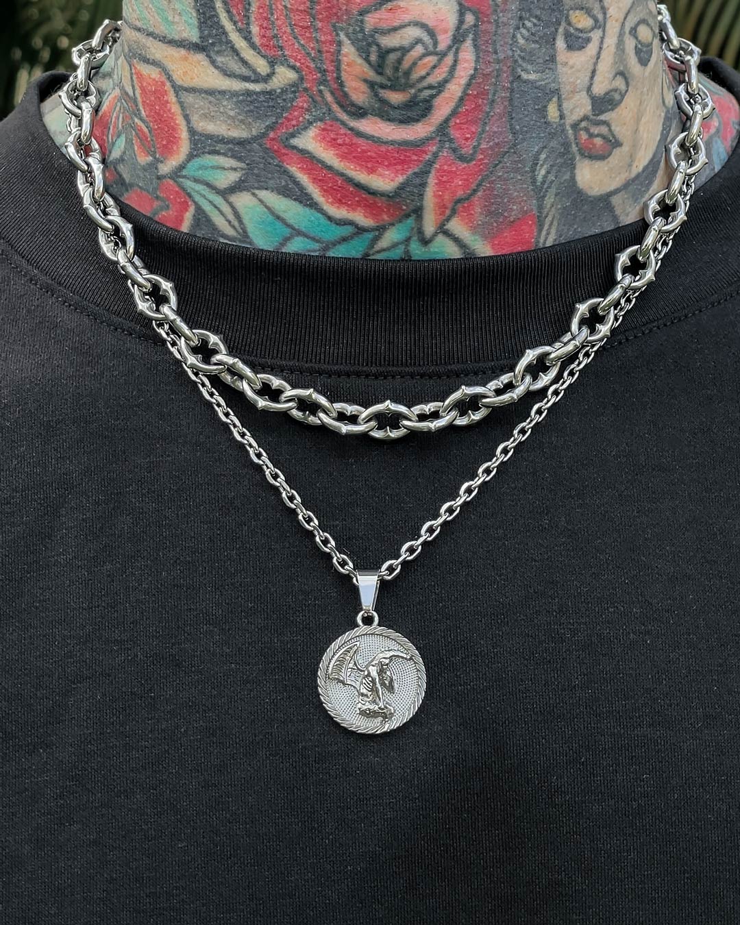 A person with colorful neck tattoos wears "The Cathedral" 9mm Spiked Chain and a finer stainless steel pendant necklace over a black shirt.