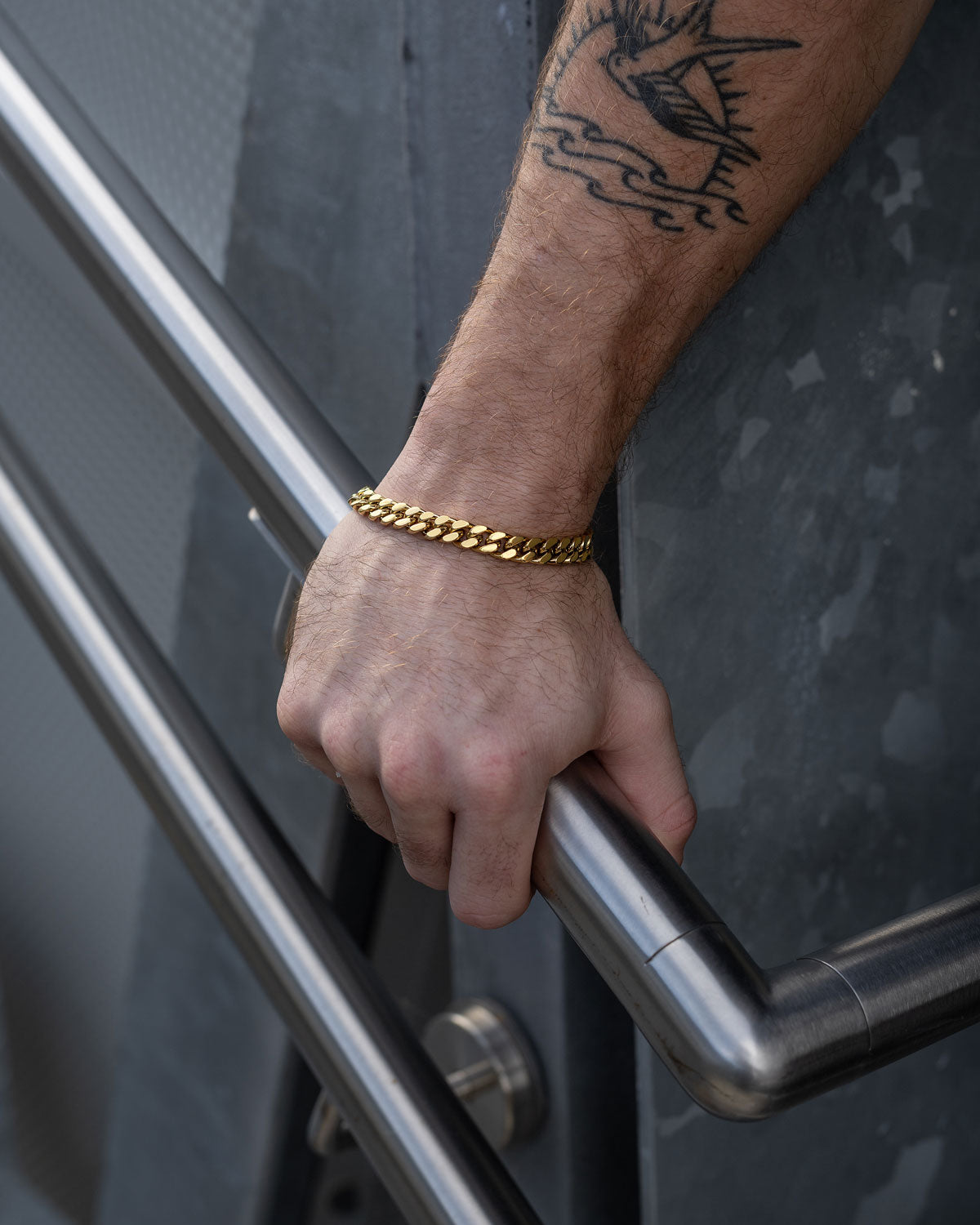 A person with a bird tattoo on their forearm wears a 9mm Cuban Bracelet (Gold) and holds a metal handrail.