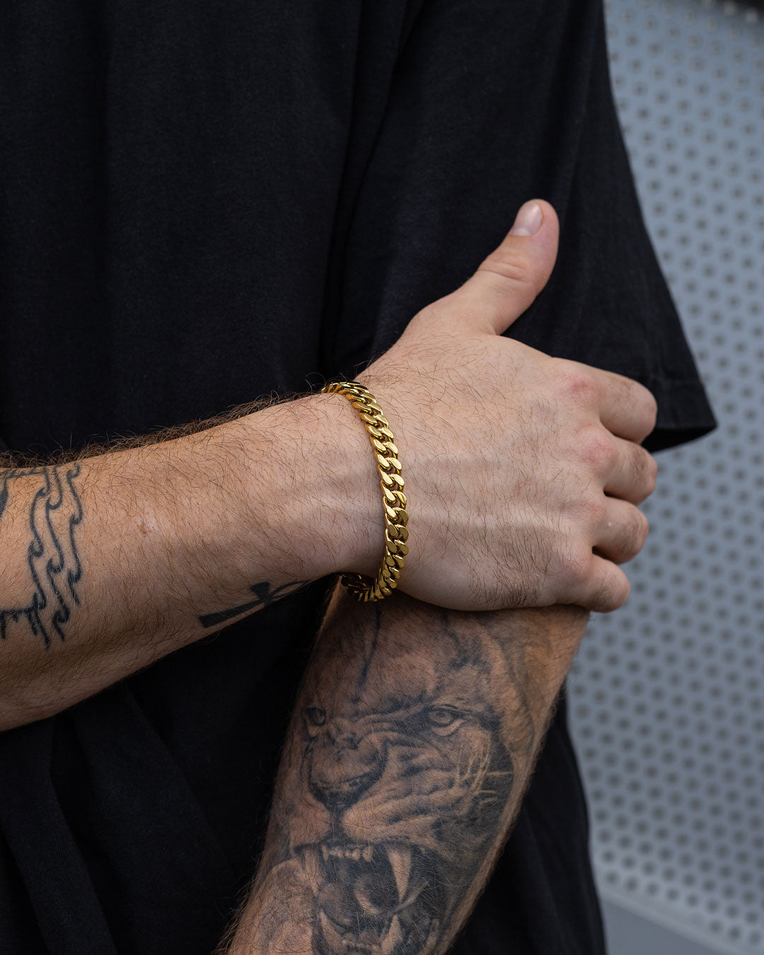 A person in a black shirt showcases their arm, featuring the 9mm Cuban Bracelet (Gold) and various tattoos—including a detailed lion face and abstract designs—set against a gray, dotted background.