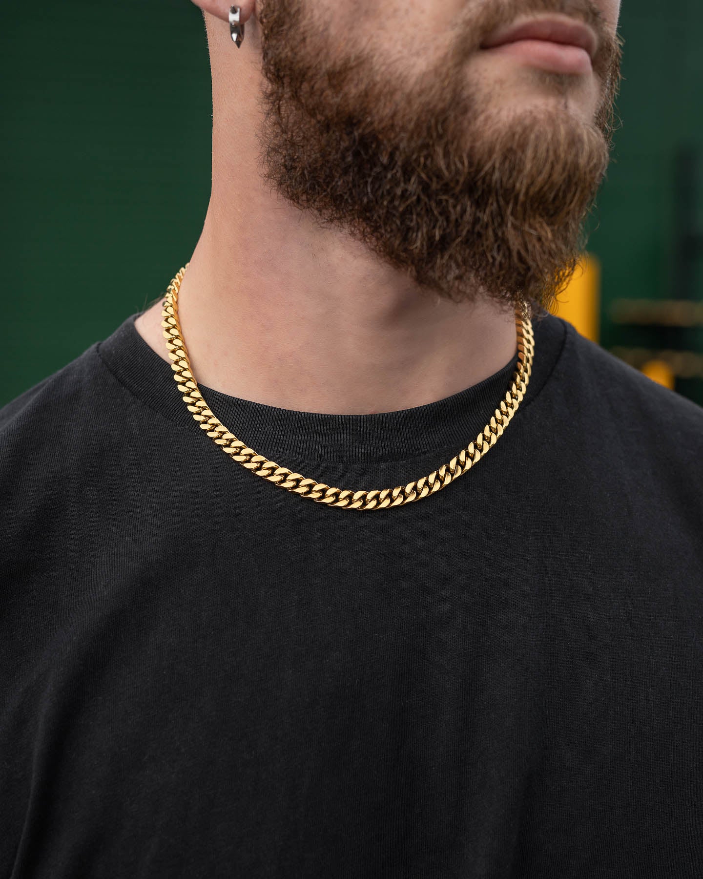 A bearded man wearing a plain black shirt and the 9mm Cuban Chain (Gold), photographed from the shoulders up against a blurred green and yellow background.