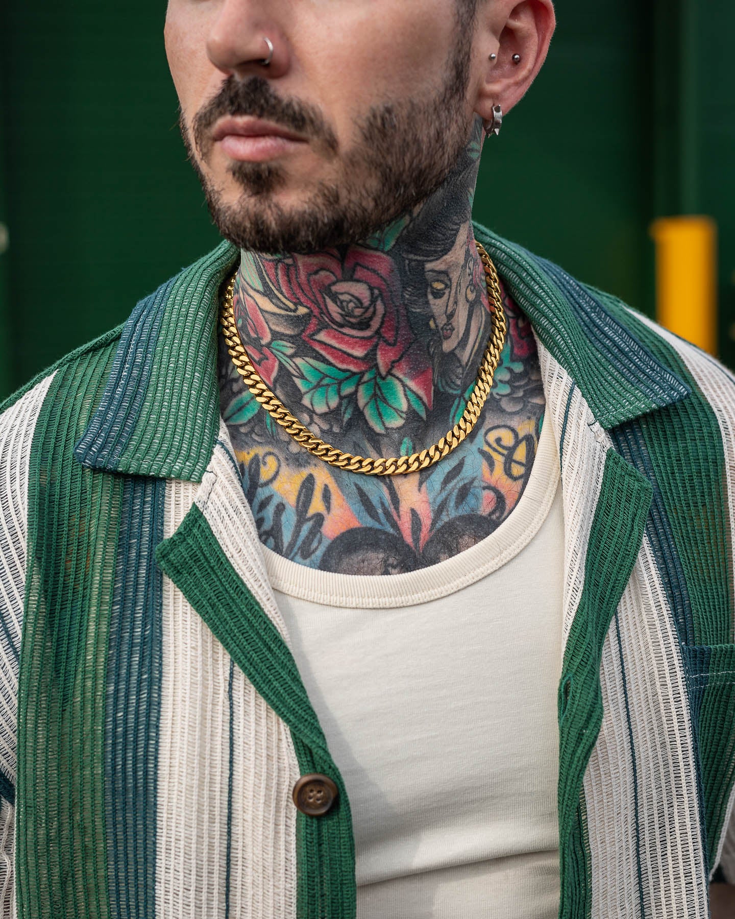 Someone wearing a green and white striped shirt over a white tank top displays colorful neck and chest tattoos, an 18k gold plated 9mm Cuban Chain (Gold), multiple ear piercings, and a nose ring.