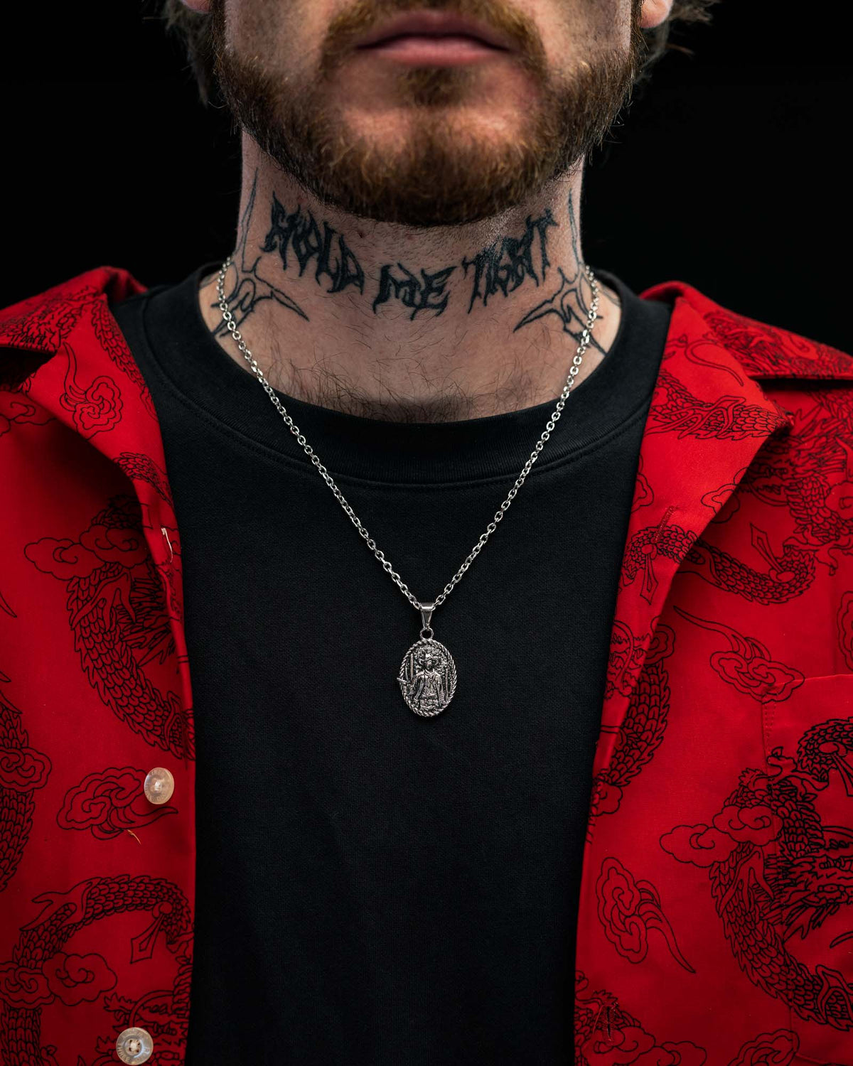 A person wearing a red shirt with black dragon patterns, a black t-shirt, the STATEMENT “Big Iron” Pendant on a silver necklace, and a large tattoo on their neck reading LAND OF TRUST.