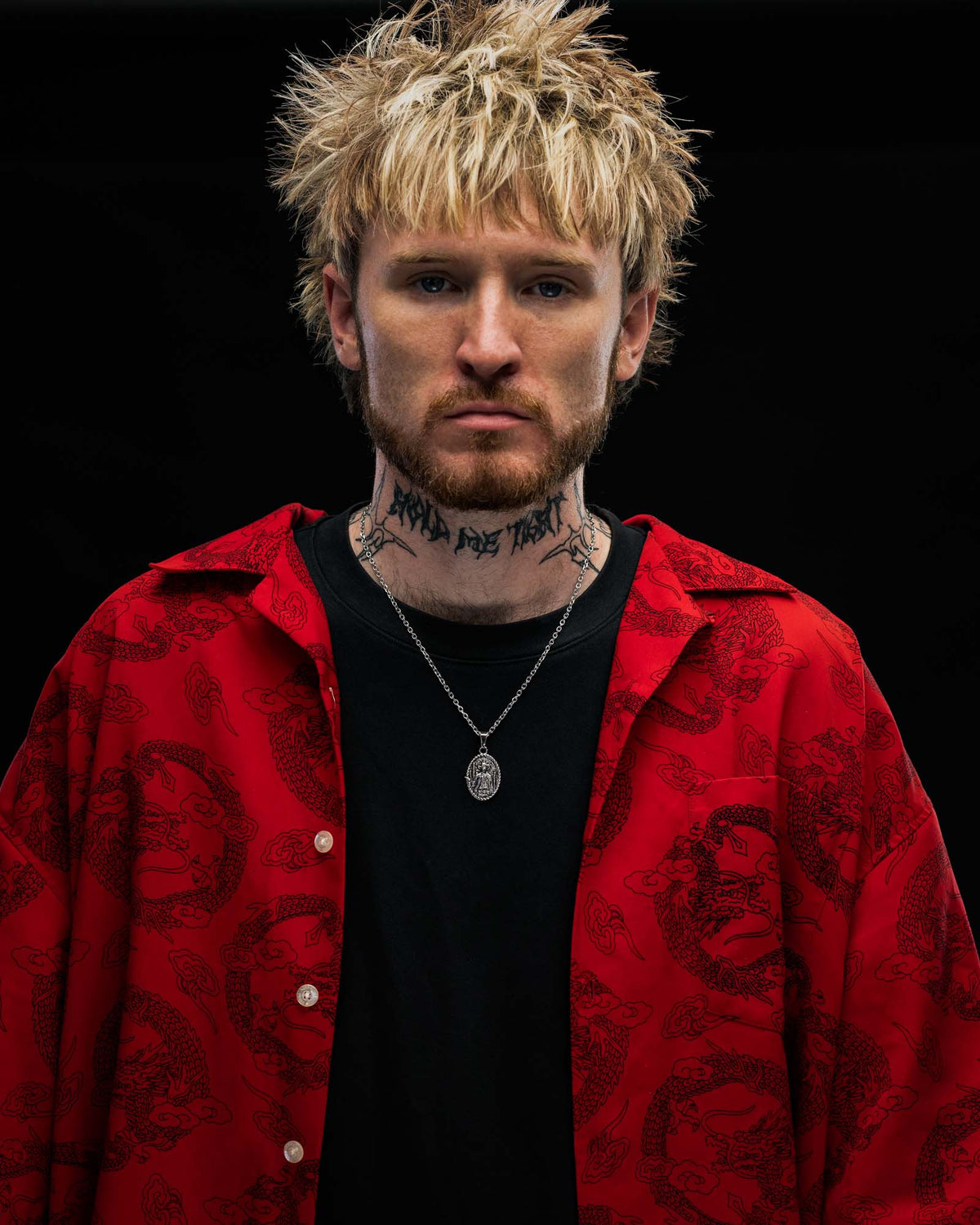 A person with messy blond hair, a beard, and neck tattoos stands against a dark background, wearing a red patterned jacket over a black shirt and showcasing the STATEMENT “Big Iron” Pendant necklace.