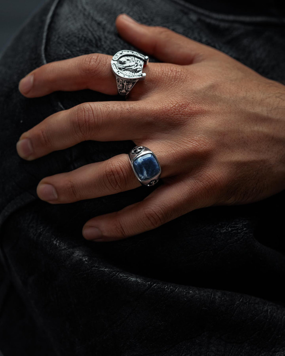A close-up of a hand resting on black leather, adorned with two ornate rings: the Statement Collective Sodalite Stone Ring featuring a vivid blue stone on the middle finger, and an intricately designed stainless steel ring—evoking the aesthetic of graveyard jewelry—on the index finger.