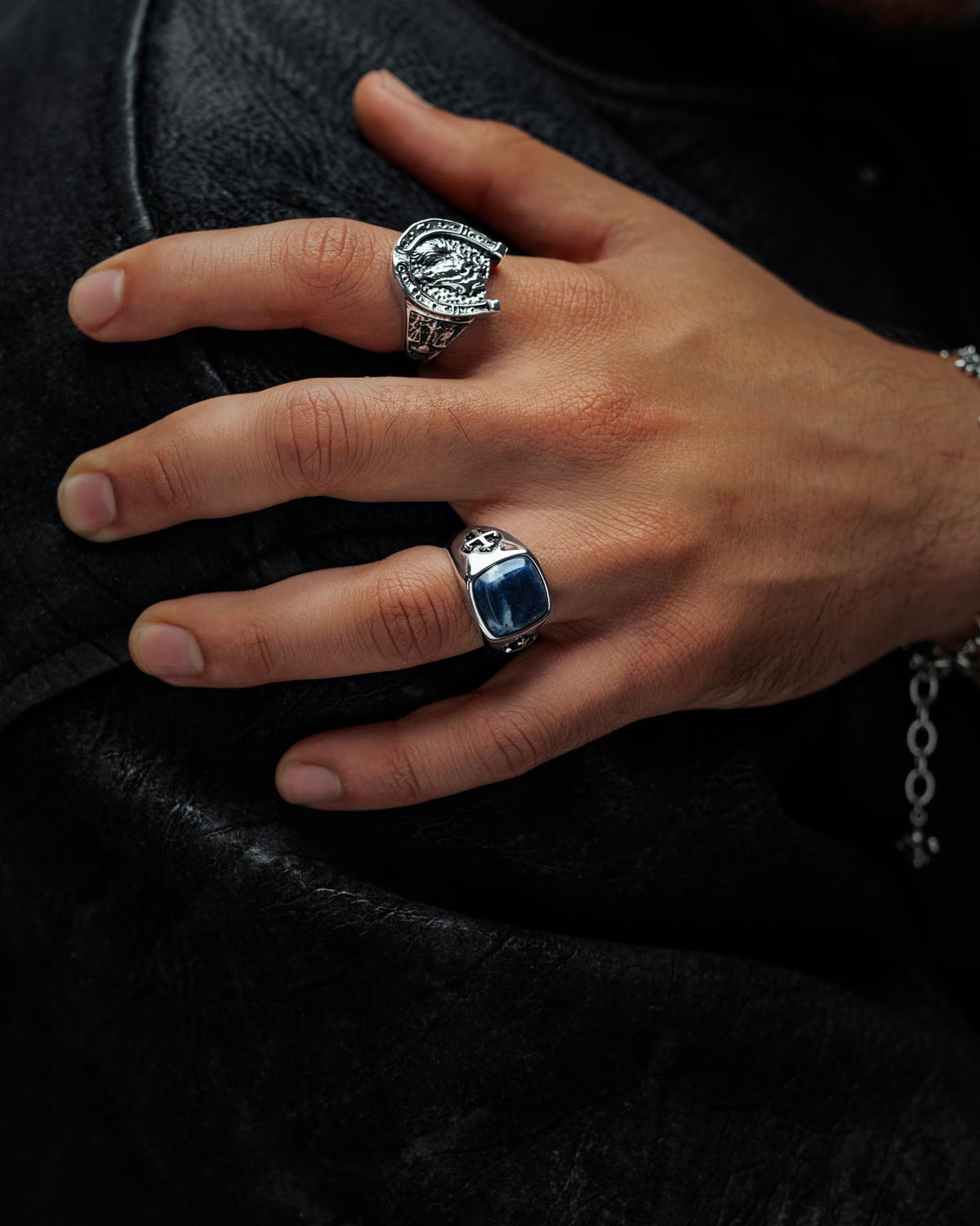 A close-up of a hand resting on dark fabric, adorned with two silver rings—including the Statement Collective Sodalite Stone Ring, which features a blue stone and cross—alongside a macabre graveyard-inspired ring and a sleek silver chain bracelet.