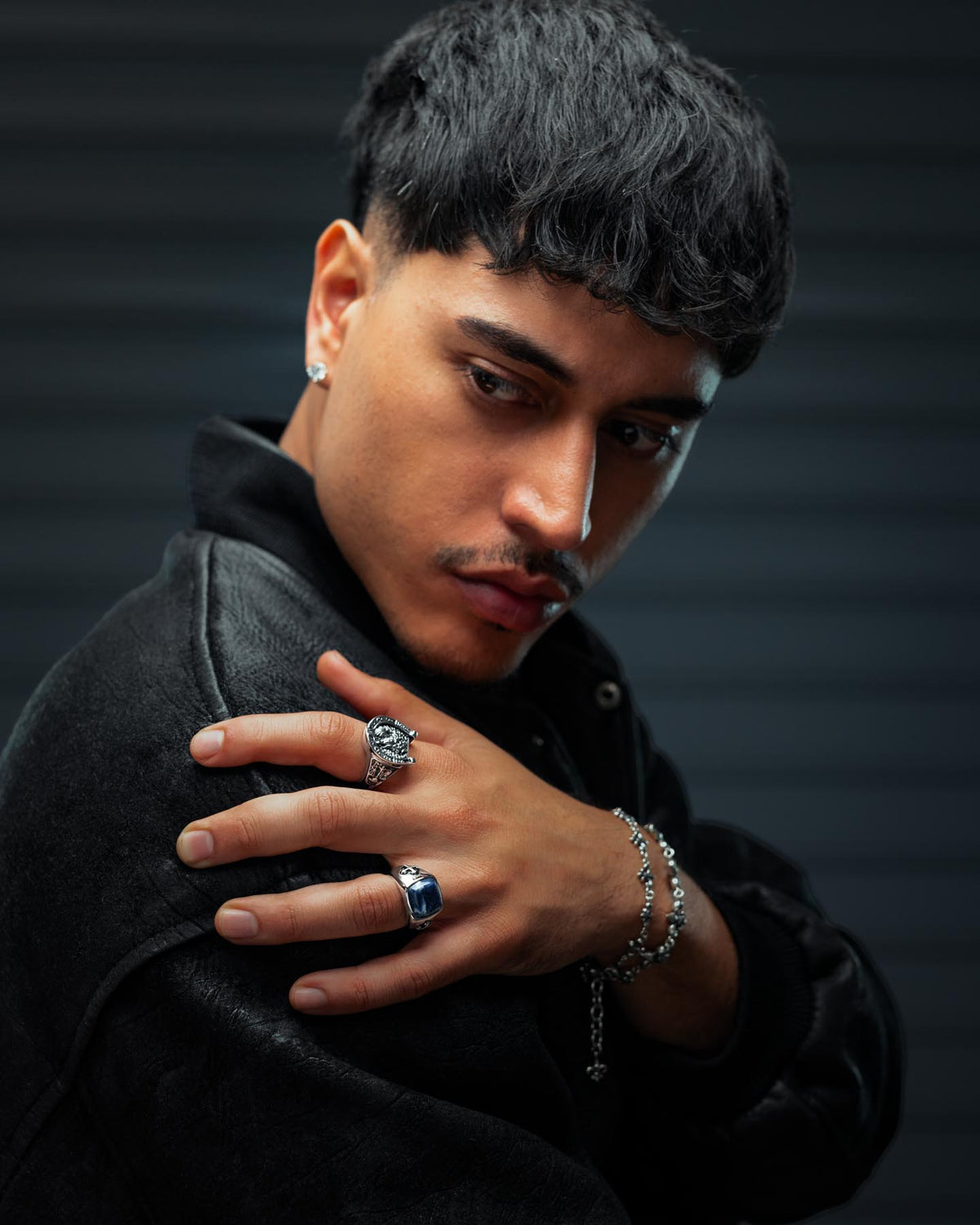 A man with short black hair and earrings looks down over his shoulder, wearing a dark jacket and showcasing the striking Sodalite Stone Ring by Statement Collective on his left hand, paired with a bracelet. The softly blurred background features horizontal lines.