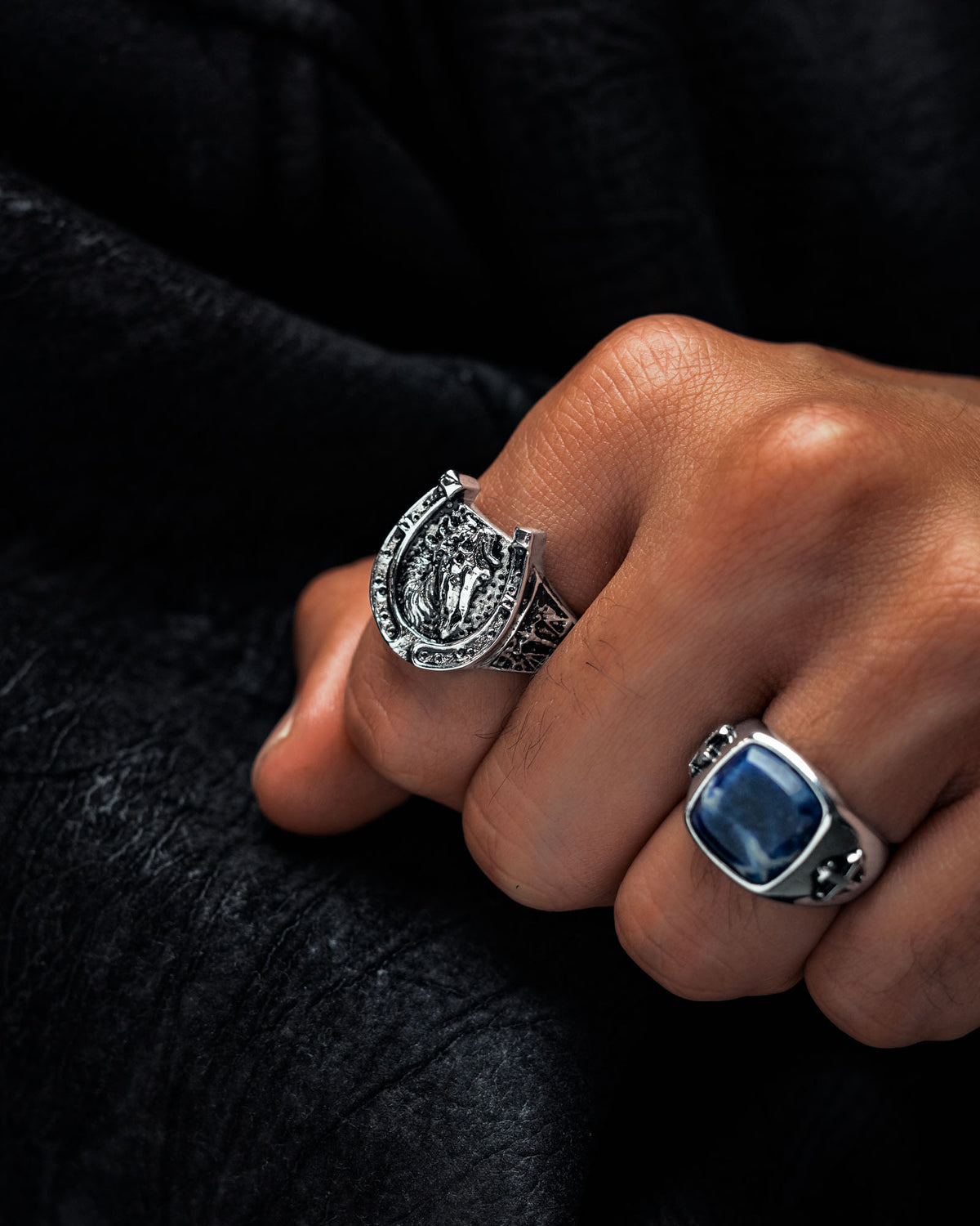A hand wearing two silver rings—a Statement Collective Bronco Ring with an engraved horseshoe design and another adorned with a large blue stone reminiscent of macabre jewellery—rests against a black textured surface.