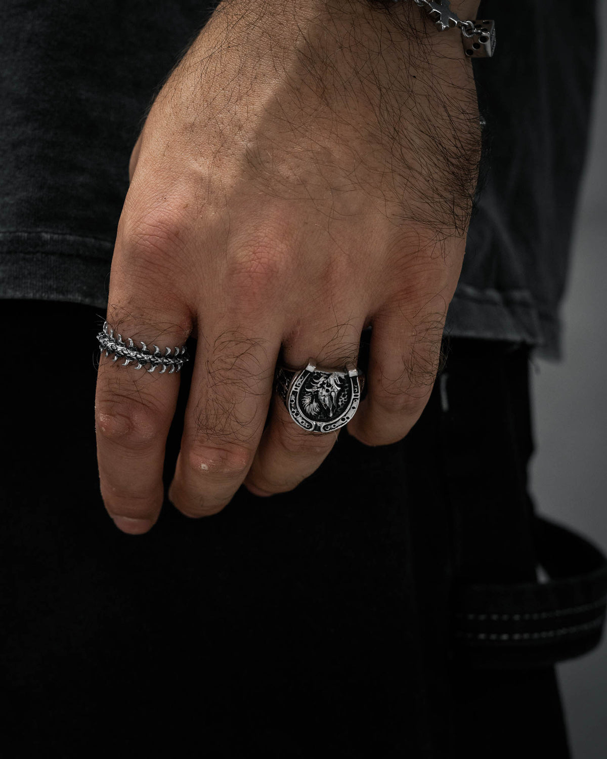 A close-up of a person’s hand wearing two silver rings, including Statement Collective’s Bronco Ring with its detailed lion’s head design alongside a chain-patterned band, showcases striking macabre jewellery against a dark background.