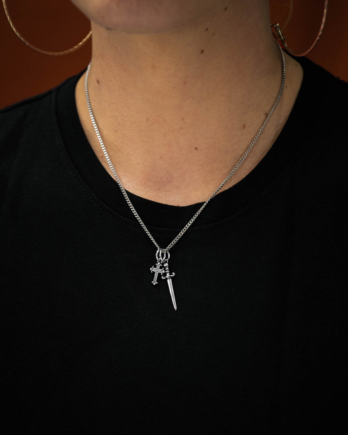 Wearing the Statement Collective Cross & Dagger Necklace—a stainless steel piece featuring both a dagger and cross pendant—a person in a black shirt perfectly captures the allure of dark romance.