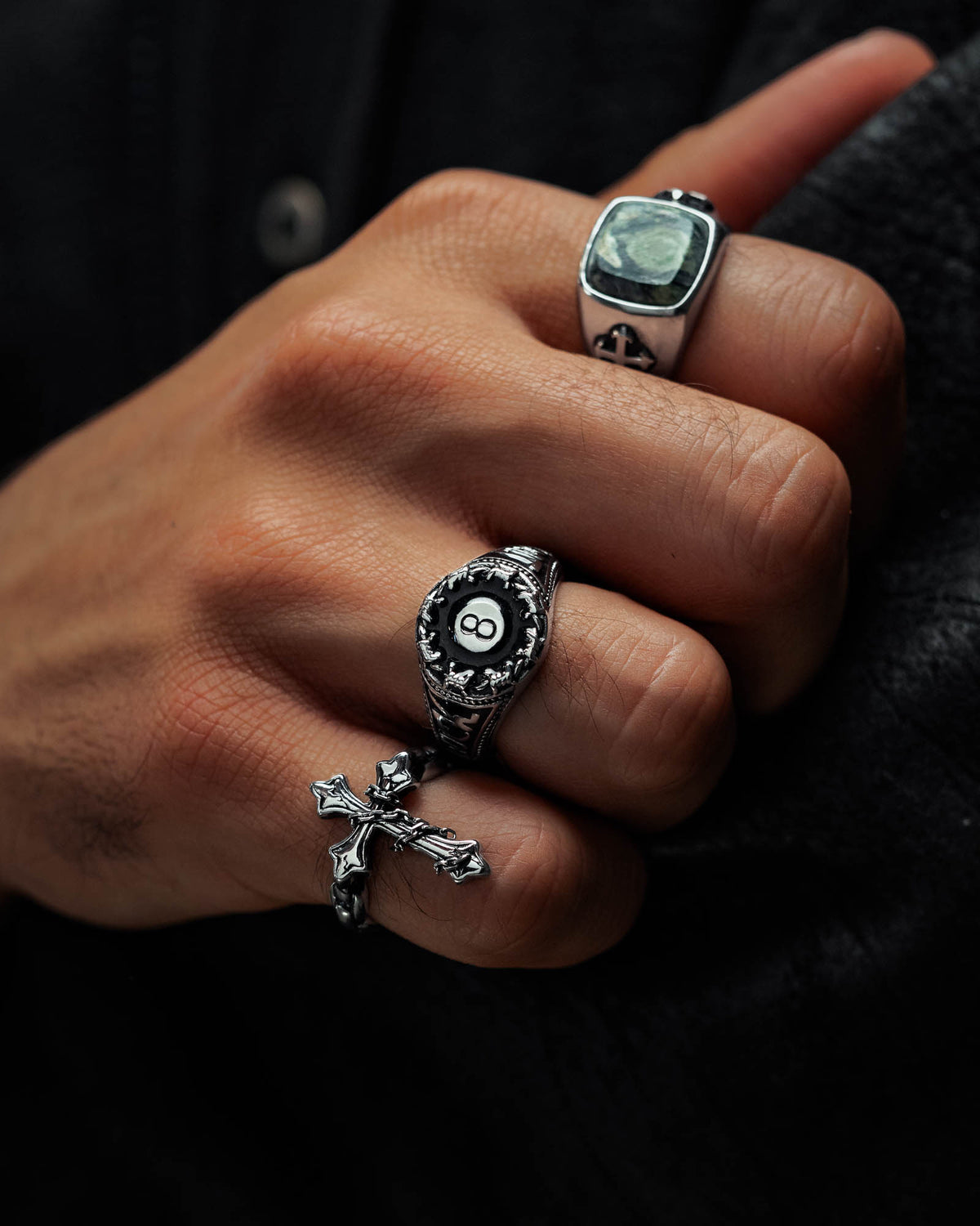 A hand adorned with three silver rings—one featuring a cross, one with an 8-ball motif, and the striking Lucky Shot Ring by Statement Collective, showcasing a square green stone reminiscent of a gravestone. The person is dressed in a dark textured jacket.