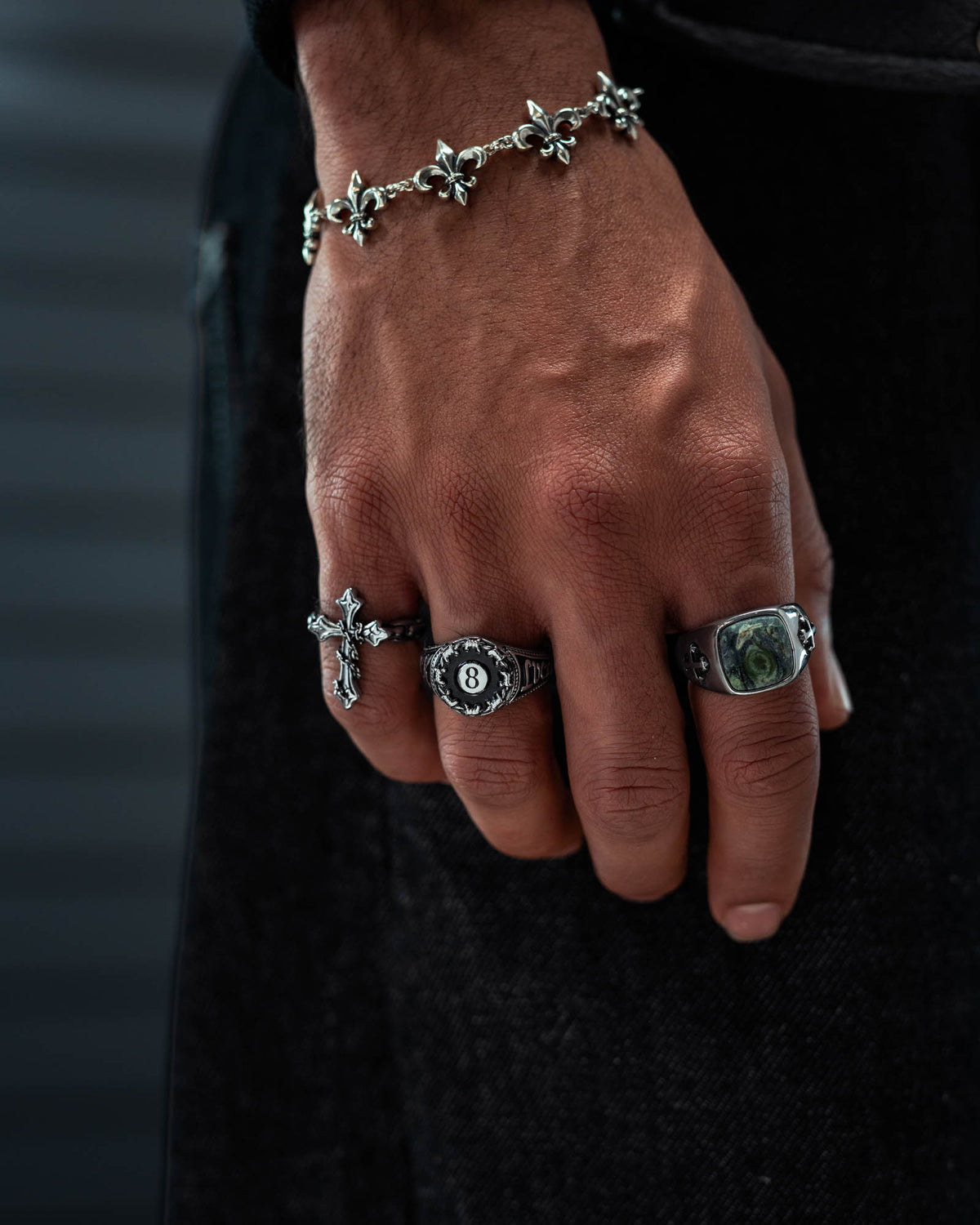 A close-up of a hand adorned with several silver rings from Statement Collective, including the Lucky Shot Ring, a cross ring, an 8 ball design ring, and a macabre graveyard-themed piece. The look is complemented by a bracelet featuring fleur-de-lis charms, all set against a dark background.