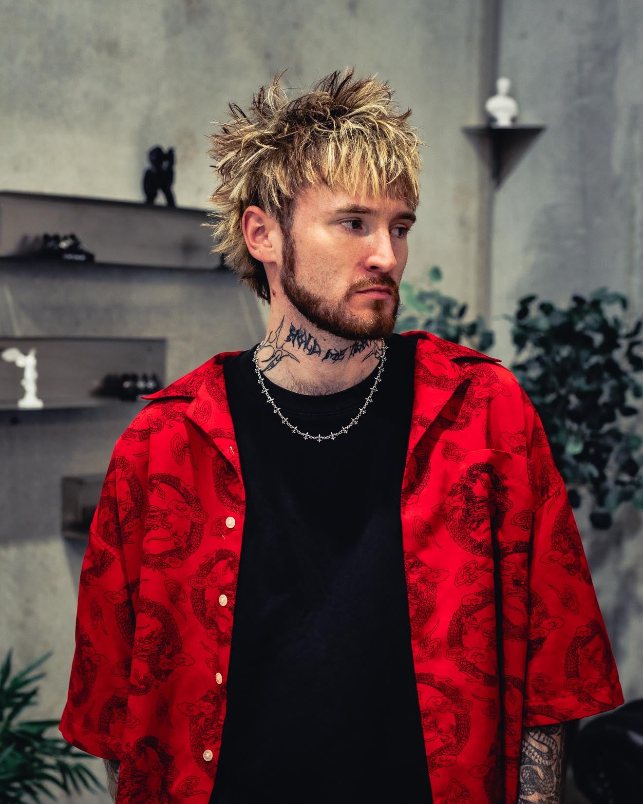 A man with spiky blond hair and neck tattoos, dressed in a black shirt layered with a red patterned button-up, stands indoors near shelves adorned with chess pieces and plants. He looks to the side with a neutral expression, wearing the Statement Collective 925 Sterling Silver “Fleur De Lis” 10mm Chain around his neck.