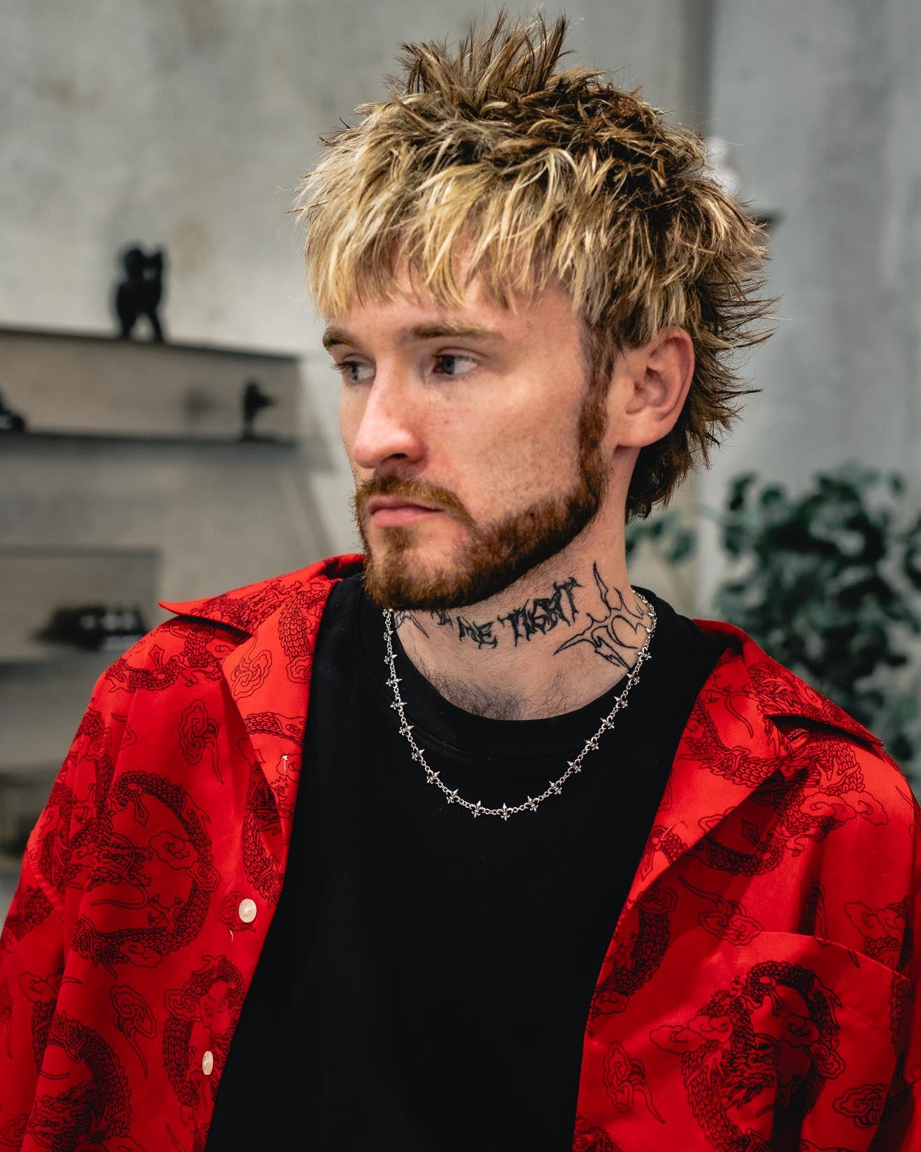 A man with shaggy blond hair, a beard, and neck tattoos wears the Statement Collective 925 Sterling Silver “Fleur De Lis” 10mm Chain over a red patterned shirt and black t-shirt. He gazes to the side, with shelves and a leafy plant visible in the softly blurred background.