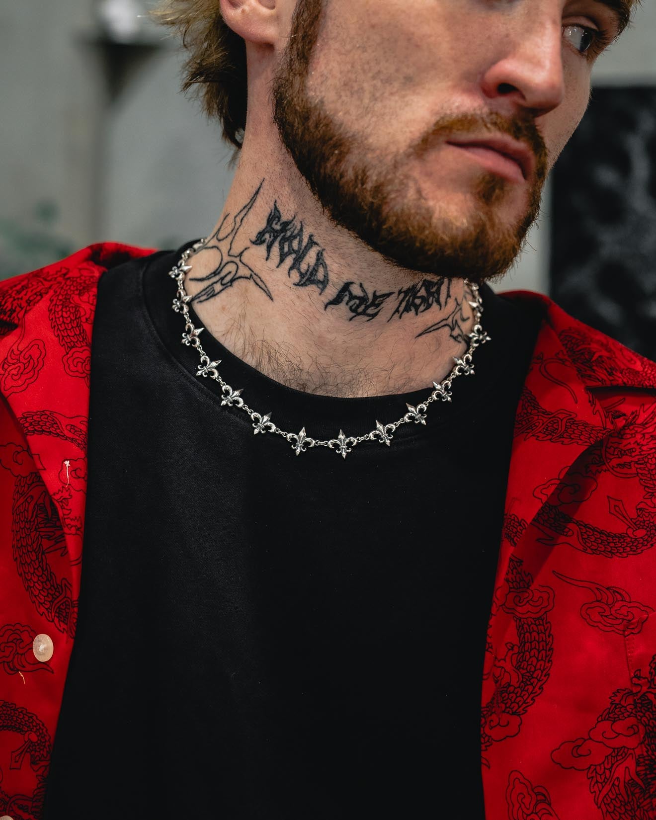 A man with a beard and neck tattoo wears the Statement Collective 925 Sterling Silver “Fleur De Lis” 15mm Chain, paired with a black shirt and a red patterned jacket. The image focuses on his neck and chest, showing part of his face.