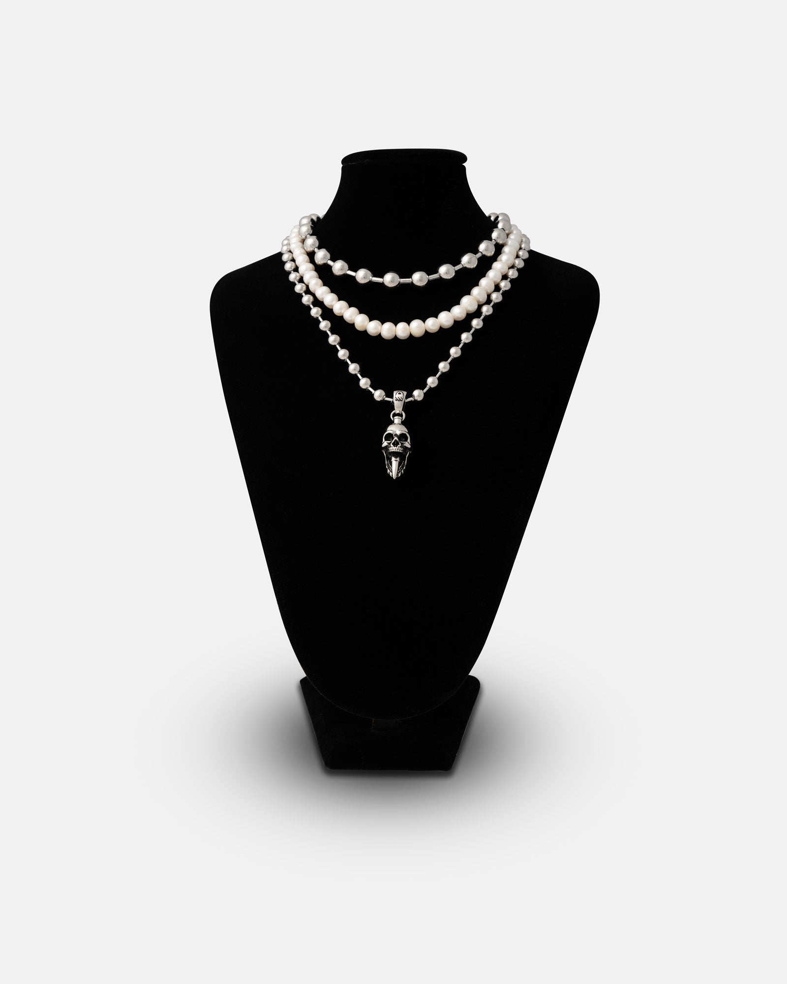 The Precious Death Stack is shown on a black jewelry bust, featuring a three-strand pearl necklace, the top chain is our No tourists choker, the middle chain is 8mm freshwater pearls and the longest chain is our 7mm ball chain with a bullet skull pendant attached with a silver skull pendant on the bottom strand, set against a plain light background.