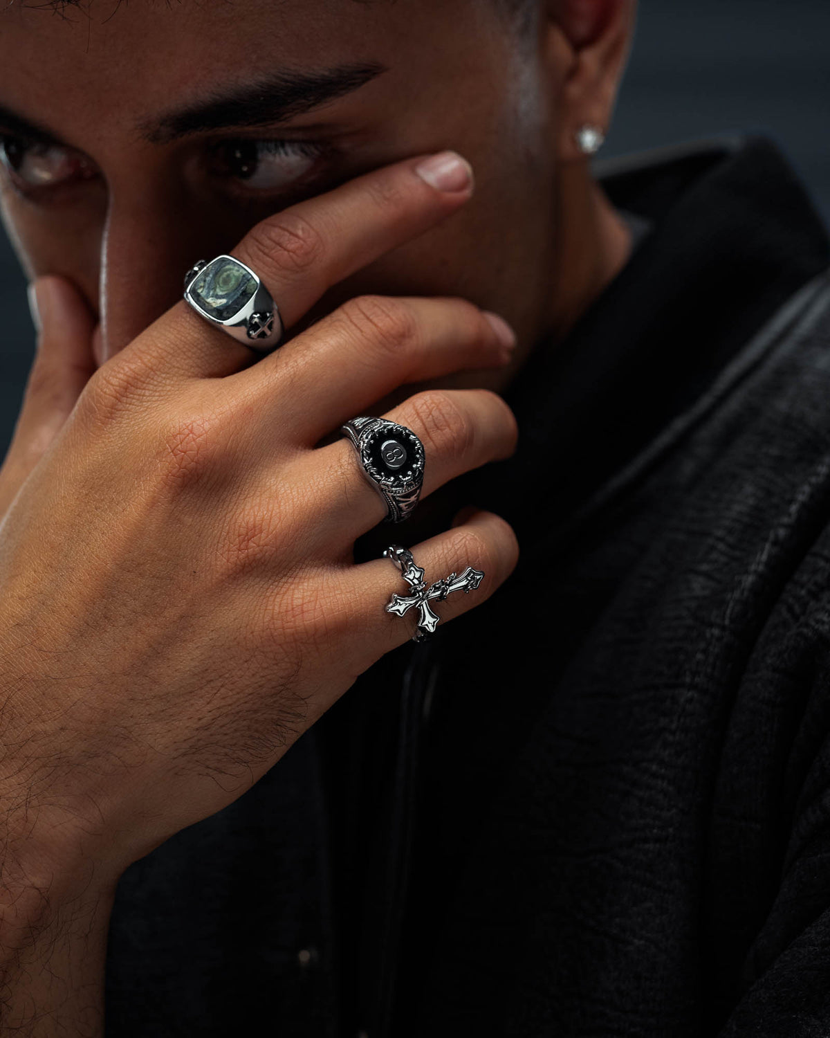 A person covers part of their face with their hand, wearing three silver rings—one adorned with a stone, one being Statement Collective’s “The Ruins” Chained Cross Ring, and one striking gravestone ring that exemplifies macabre jewelry—while dressed in a dark textured jacket.