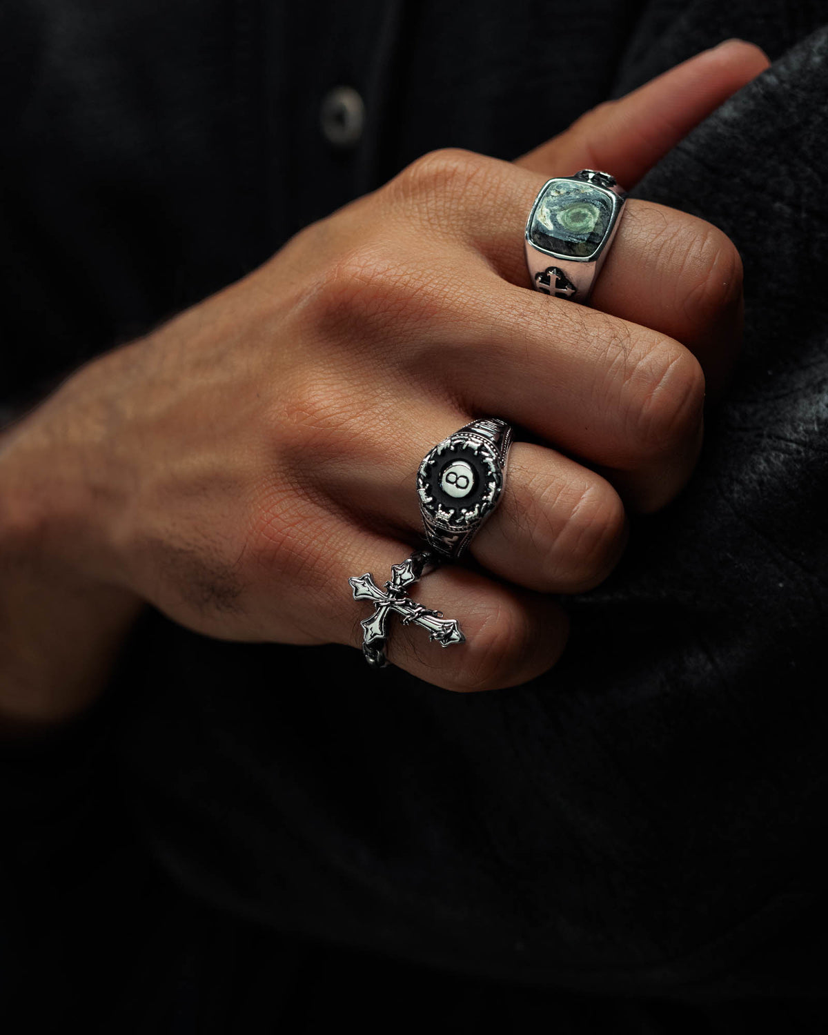A close-up of a hand wearing three silver rings: one with a green stone, one featuring an eight-ball design, and Statement Collective’s “The Ruins” Chained Cross Ring—an evocative piece that captures the allure of macabre jewelry—all set against a dark background.