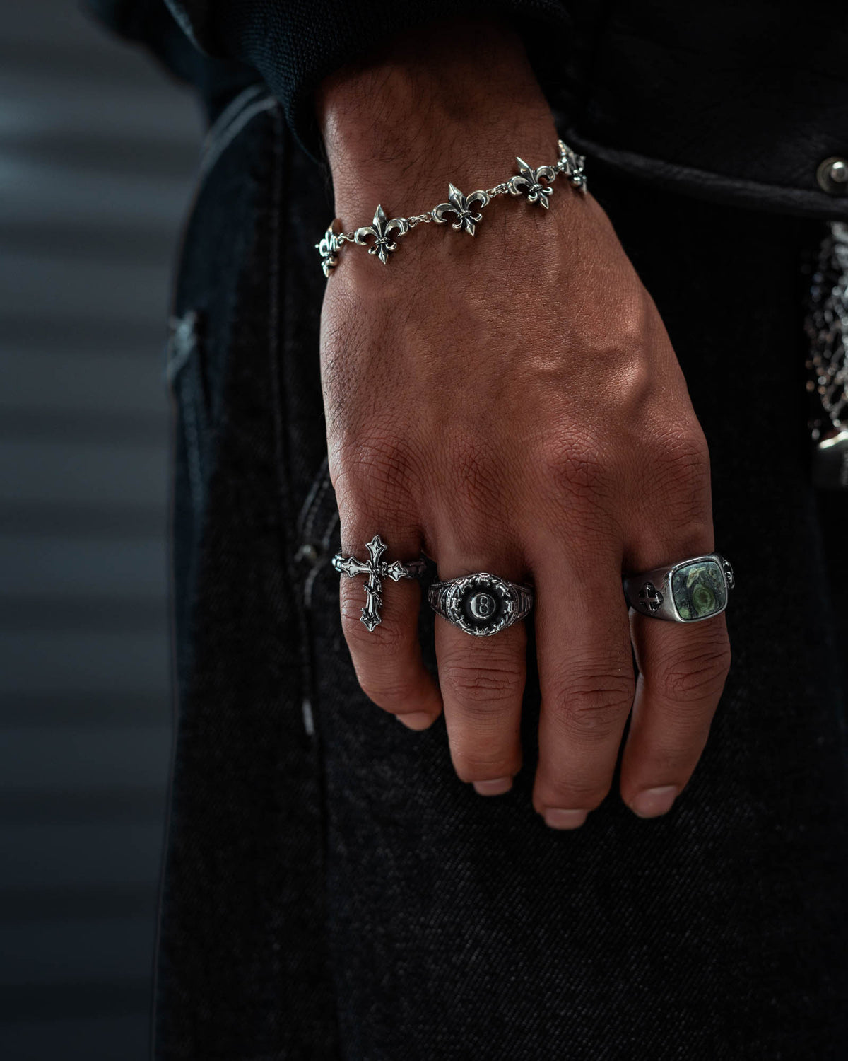 A close-up of a person’s hand wearing multiple silver rings, including Statement Collective’s “The Ruins” Chained Cross Ring and other intricately detailed designs, complemented by a bold gravestone ring that introduces a macabre touch. A silver bracelet adorned with fleur-de-lis charms completes the look, while dark clothing enhances the dramatic style.