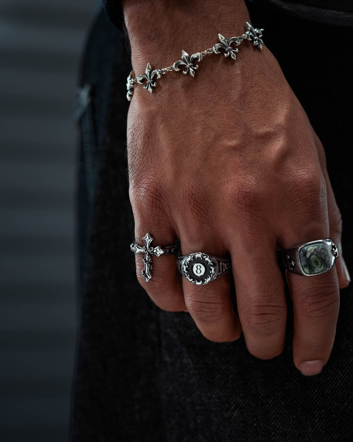 A hand adorned with three ornate silver rings—including Statement Collective’s striking “The Ruins” Chained Cross Ring—and a silver fleur-de-lis bracelet, the focus resting on the macabre jewelry against a dark, blurred background.
