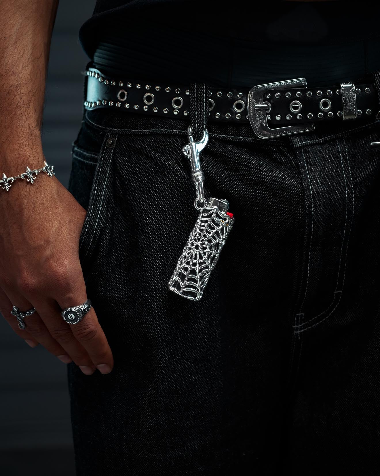 A person in dark jeans and a studded belt has the Statement Collective Spiderweb Lighter Sleeve—crafted from sterling silver—clipped to a belt loop. Their hand, decorated with rings and a bracelet, rests nearby.