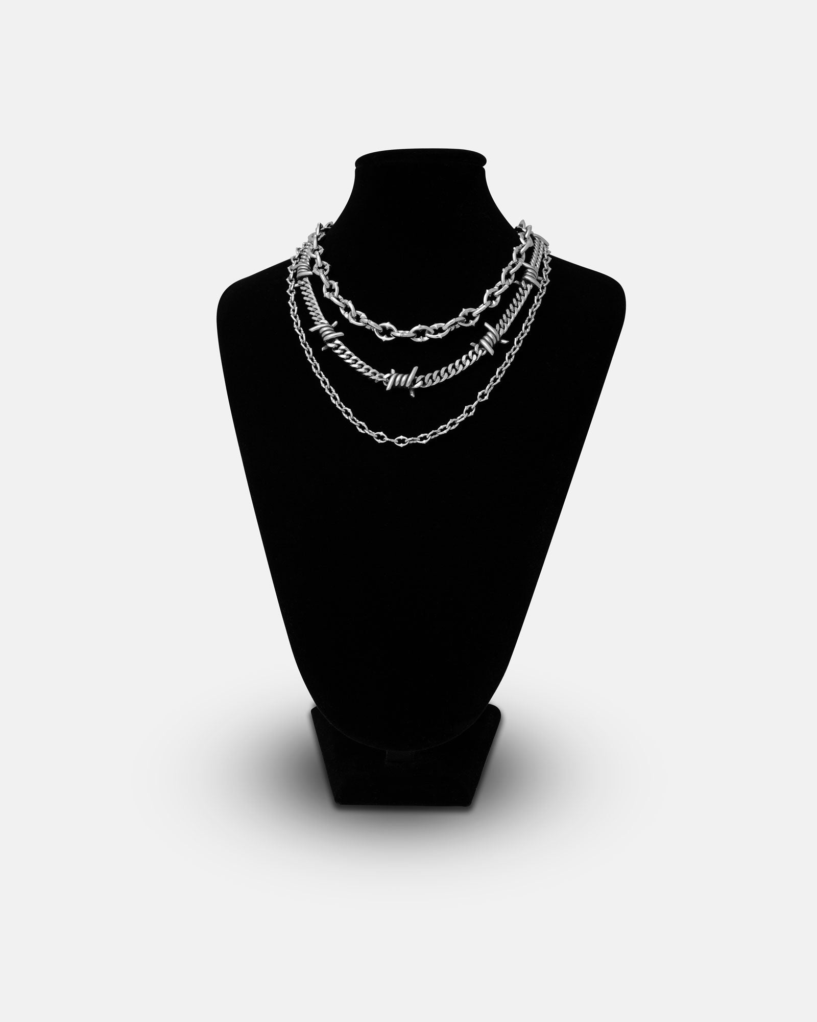A black jewelry bust showcases the Stay Safe Stack: a layered silver necklace set with three unique chain styles—barbed wire and spiked chains—displayed against a plain white background.