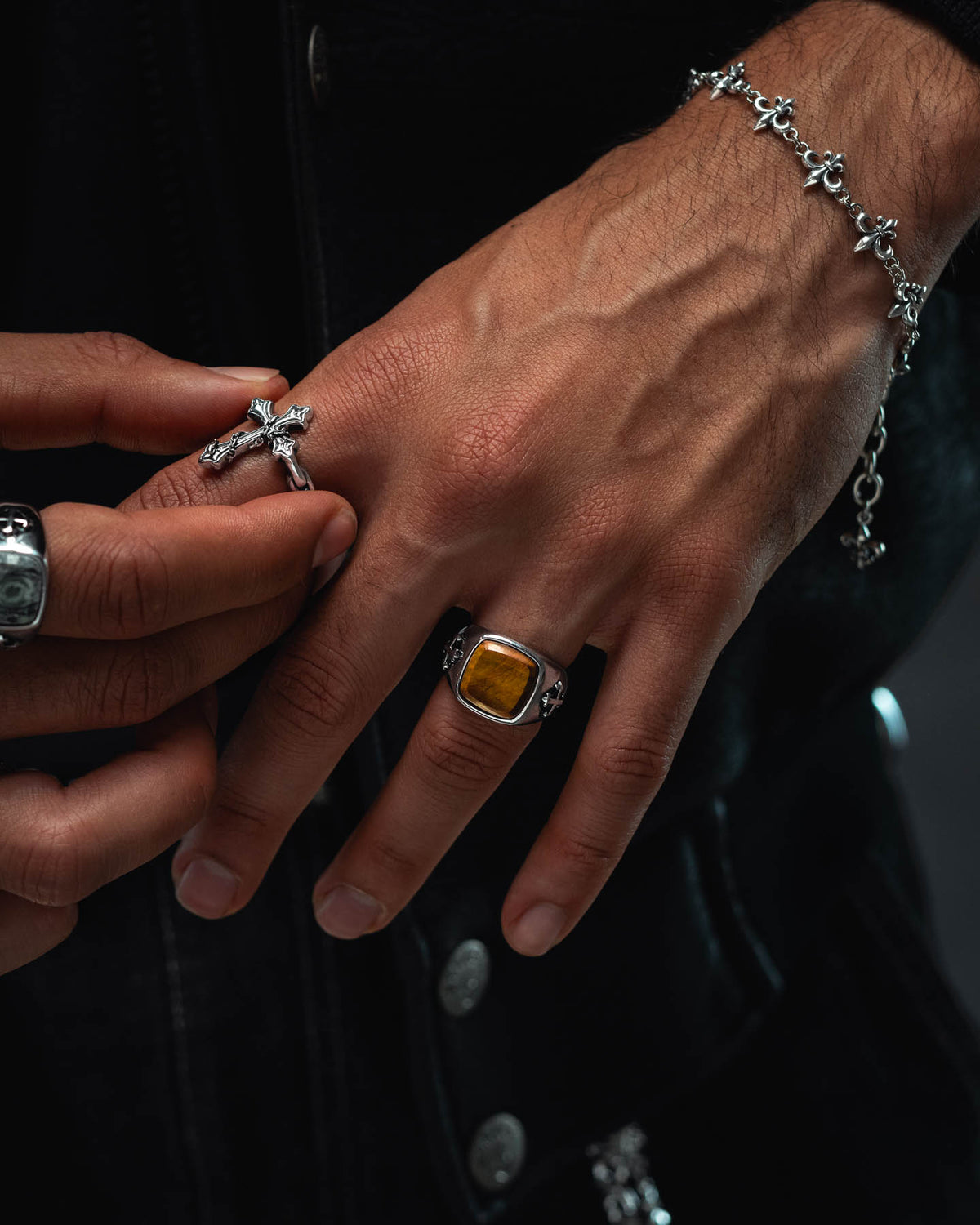 A close-up of two hands with prominent veins, adorned with silver rings—including the Statement Collective Tigers Eye Stone Ring—a cross bracelet, and holding a cross necklace. The Tigers Eye Stone Ring features a large yellow stone, complementing the collection of macabre jewellery. The person wears a dark, buttoned jacket.