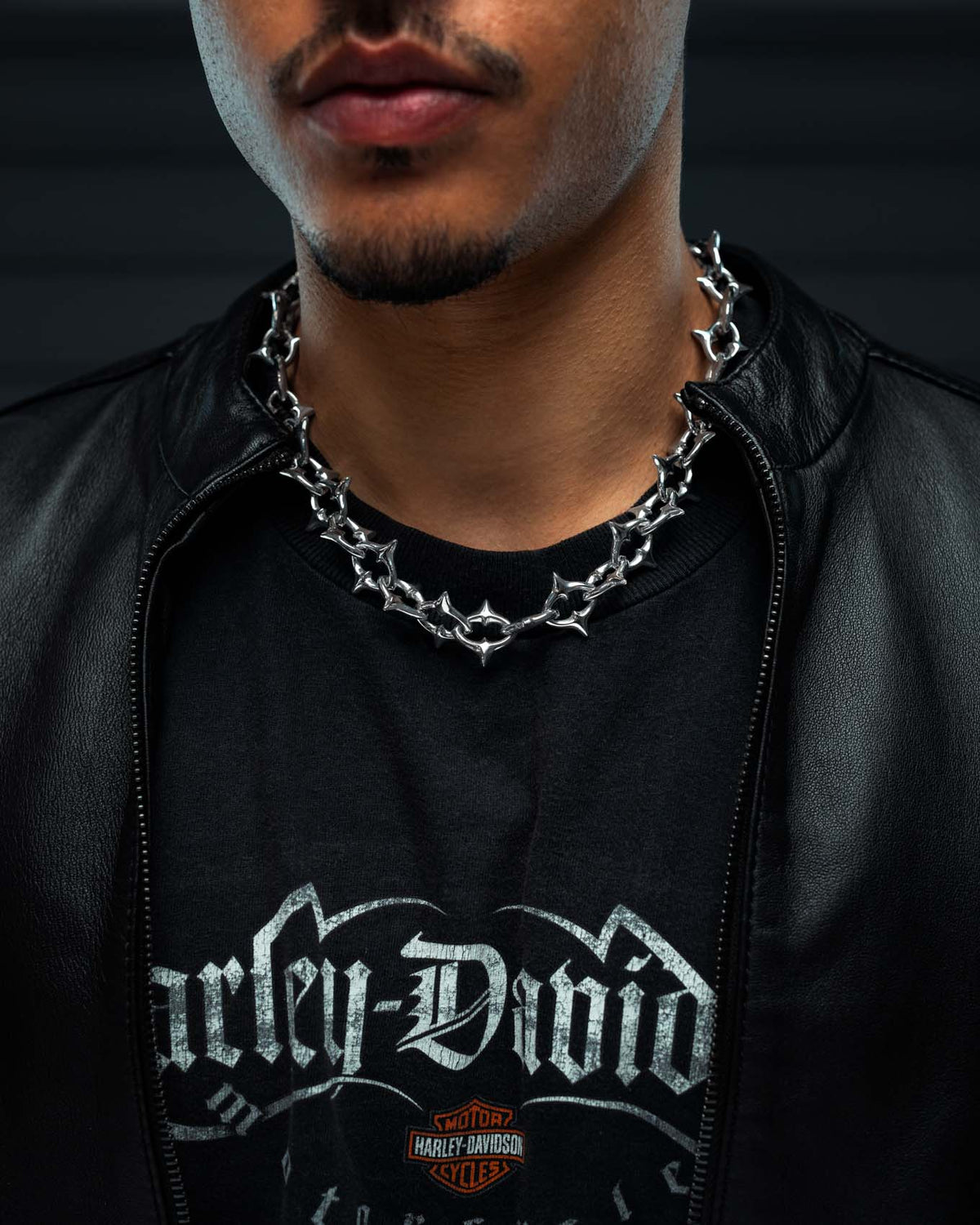 A person wearing a black Harley-Davidson t-shirt, a black leather jacket, and Statement Collective’s “The Cathedral” XL 15mm Spiked Chain necklace. Only the lower half of their face and upper torso are visible.