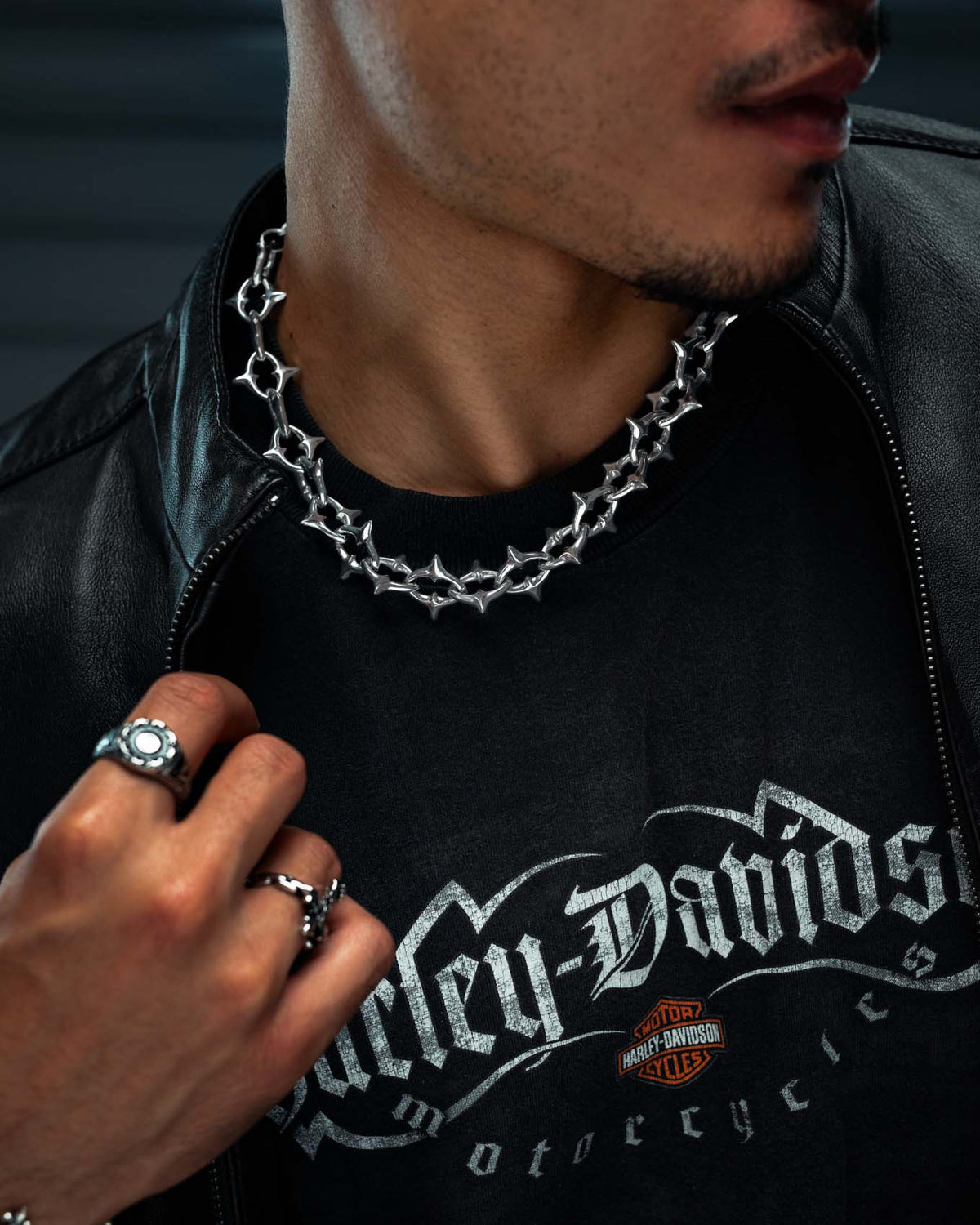 A person wearing a black Harley-Davidson T-shirt, black leather jacket, and several silver rings. Their accessories take center stage—including “The Cathedral” XL 15mm Spiked Chain by Statement Collective and other stainless steel jewelry—creating a bold, edgy look. The focus is on the striking gothic chain and statement pieces that complete their style.