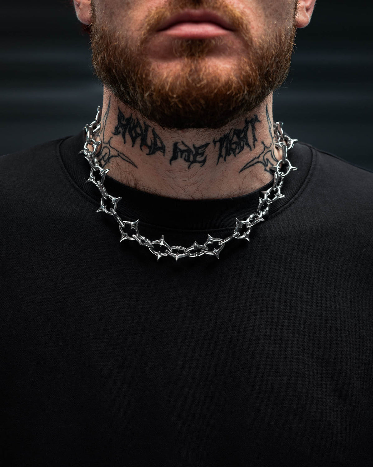 A person with a reddish beard and neck tattoo wears “The Cathedral” XL 15mm Spiked Chain by Statement Collective—a thick, gothic stainless steel necklace—paired with a plain black shirt. The background is dark and blurred.