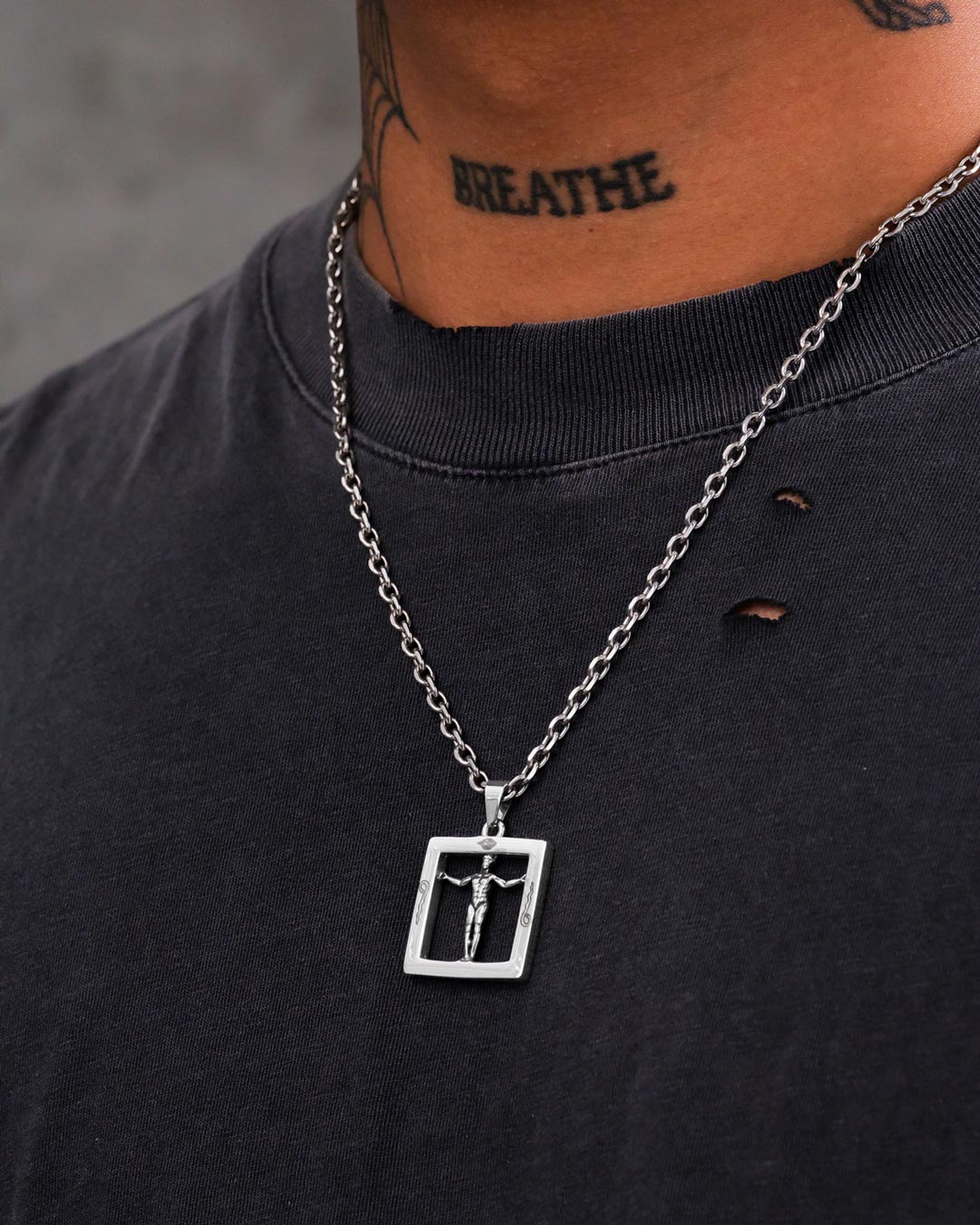 A close-up of a person in a black shirt with two small holes, wearing the Agrippa’s Square Pendant—a silver symbolic necklace—plus a “BREATHE” neck tattoo, creating a striking and mysterious jewelry aesthetic.