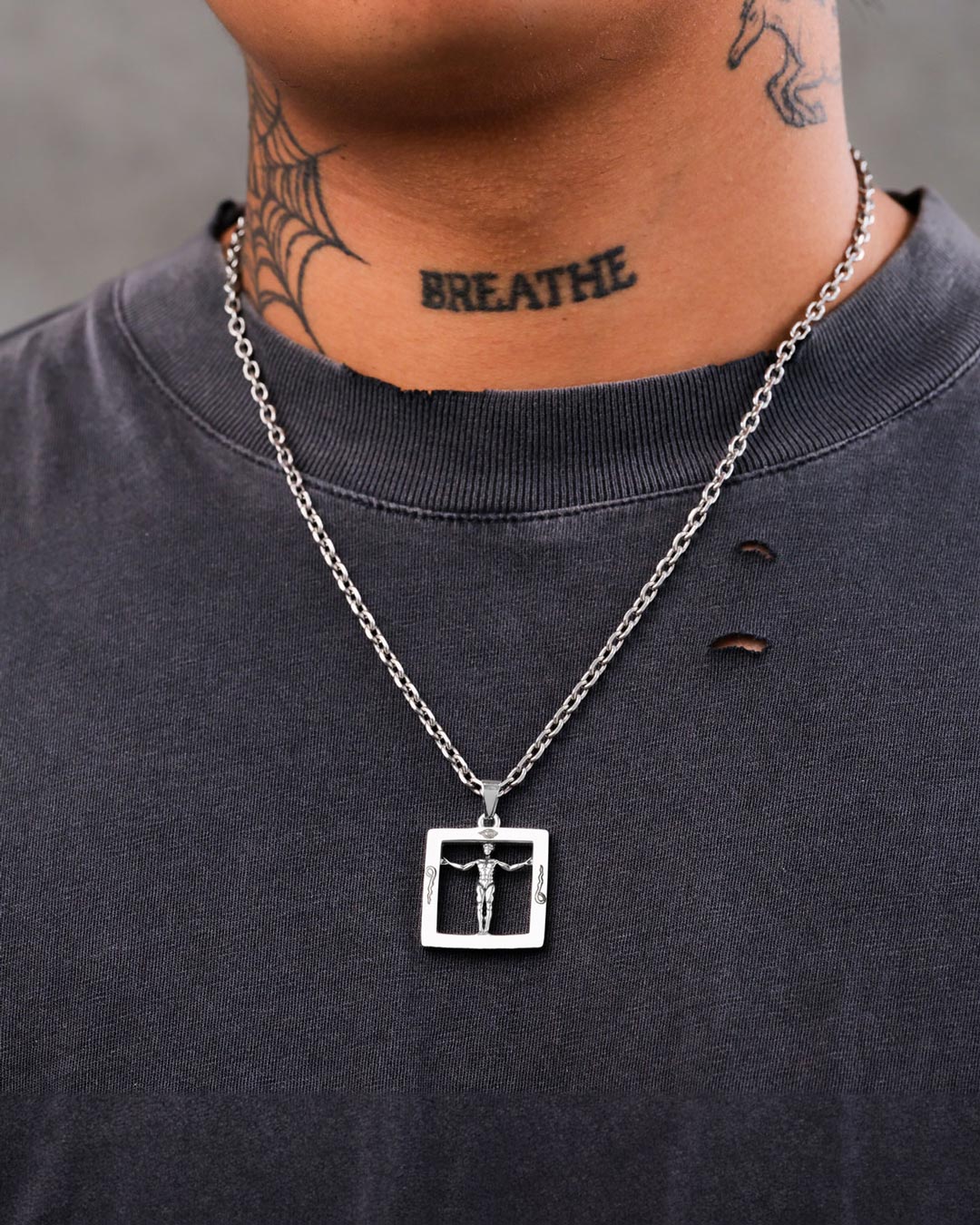 A person sports a dark, distressed t-shirt and the Agrippa’s Square Pendant—a silver occult necklace. They have a BREATHE tattoo and a spiderweb tattoo on their neck.