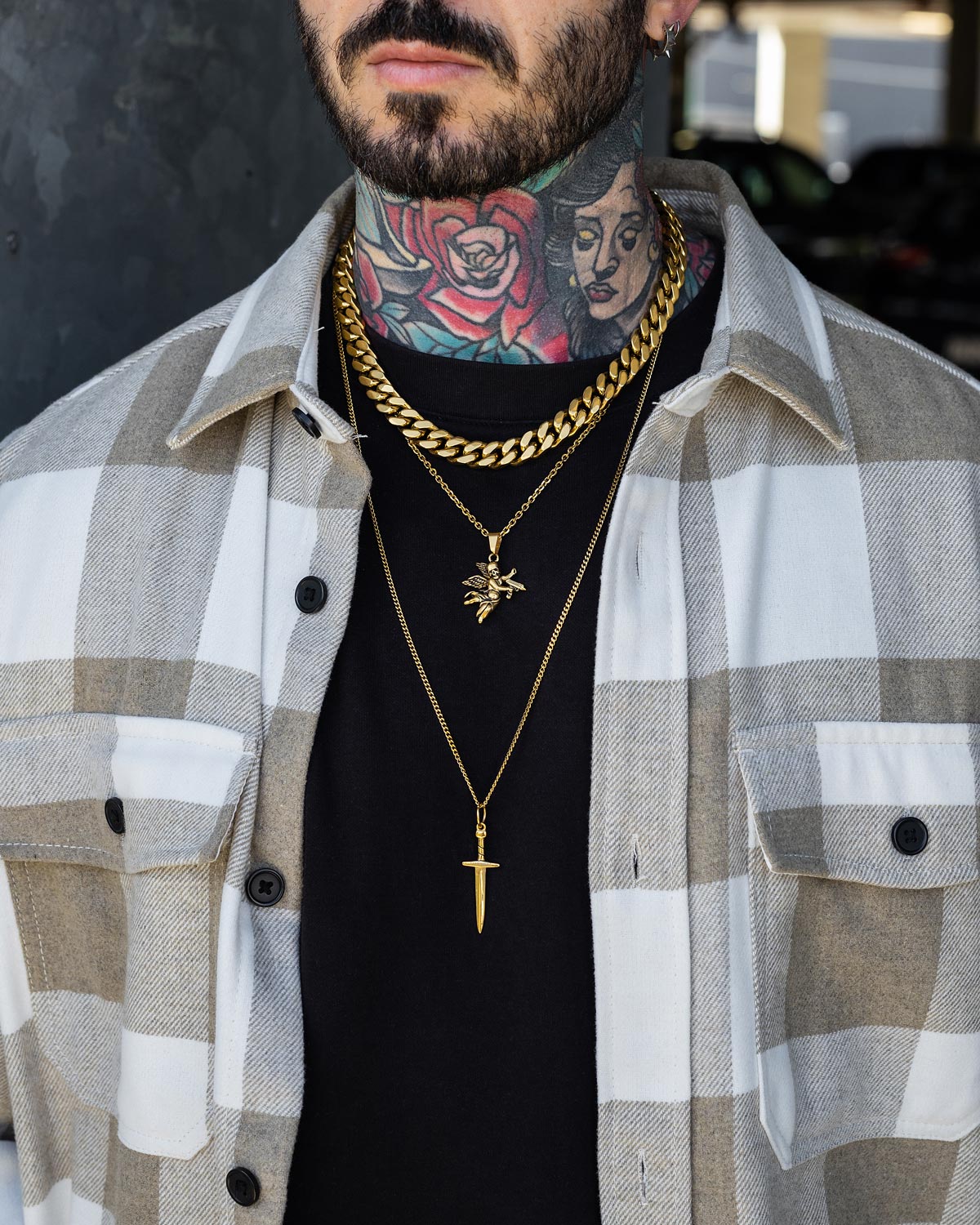 A person wears a beige and white plaid shirt over a black top, layered with the Statement Collective Angel & Dagger Stack (Gold) necklace and gold chains, showing colorful tattoos on their neck and chest.