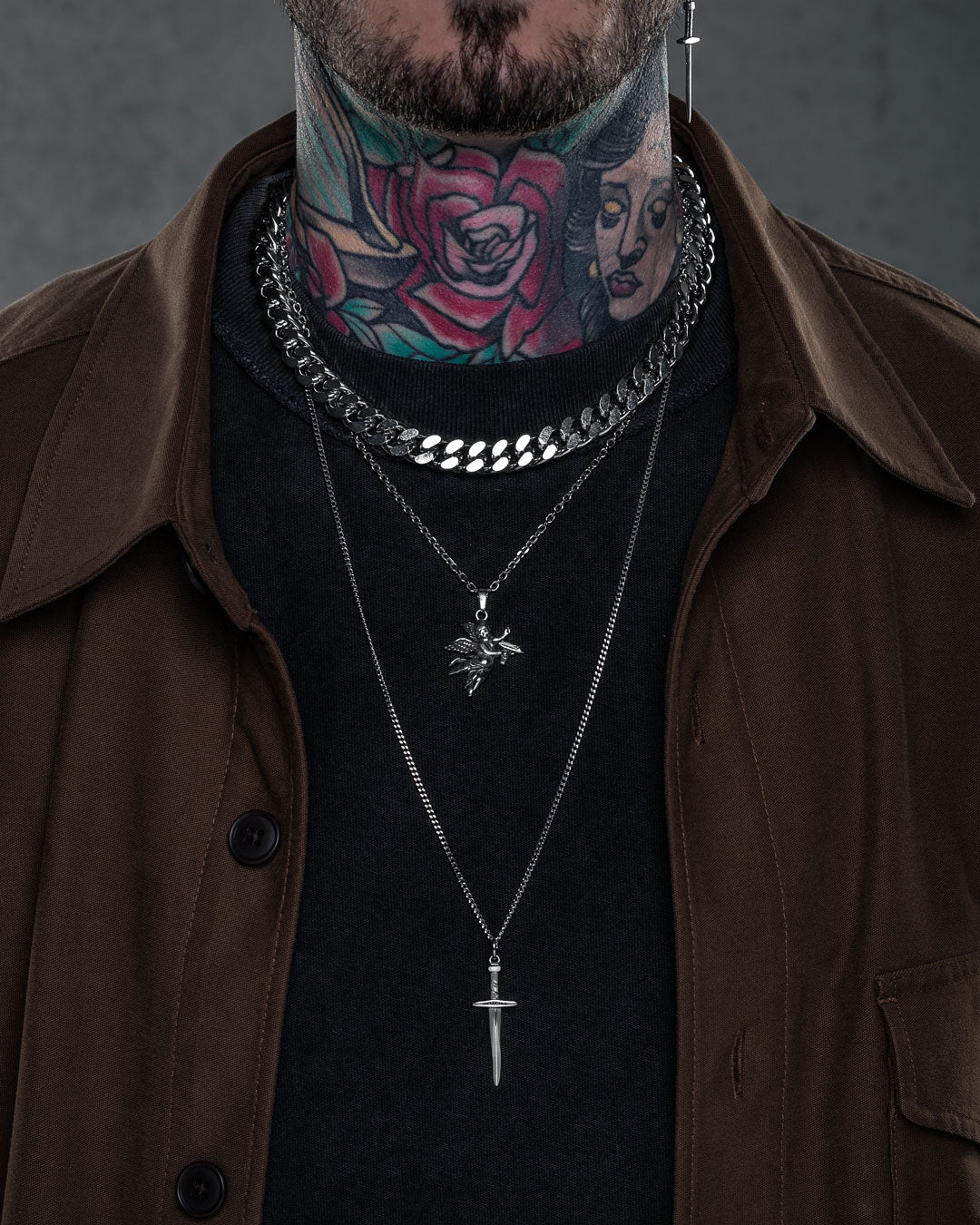 Someone wearing the Angel & Dagger Stack—layered silver Cuban link and pendants with dagger and winged figure—paired with a brown jacket, black shirt, plus bold rose and woman’s face neck tattoos.