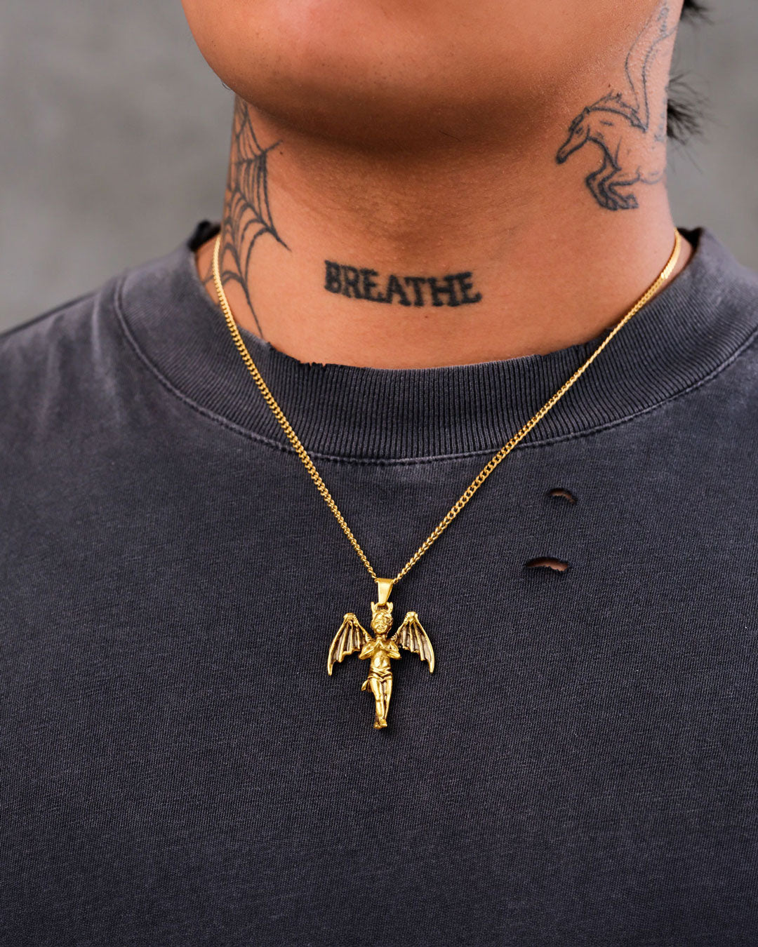 A person wearing a dark, distressed shirt and "The Wicked One" Pendant (Gold), a gothic-inspired 18k gold-plated cherub necklace. They have BREATHE and spiderweb neck tattoos, plus a bird tattoo by the ear.