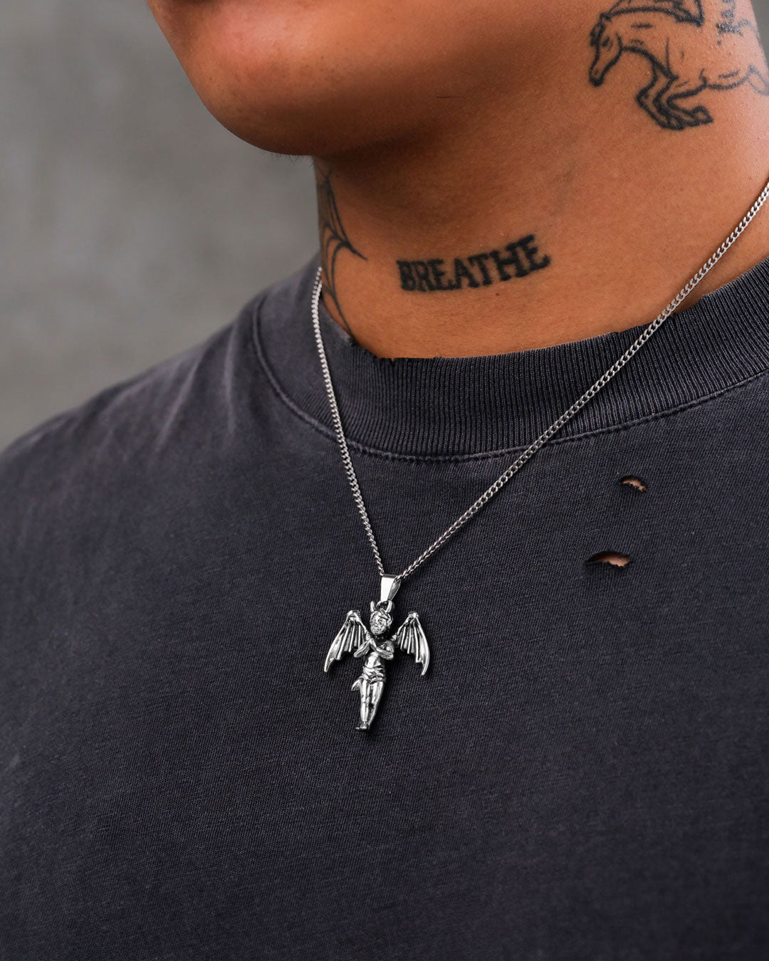 A close-up of a person wearing a black distressed t-shirt, "The Wicked One" Pendant— a stainless steel cherub necklace— and a “BREATHE” neck tattoo, set against a plain gray background.