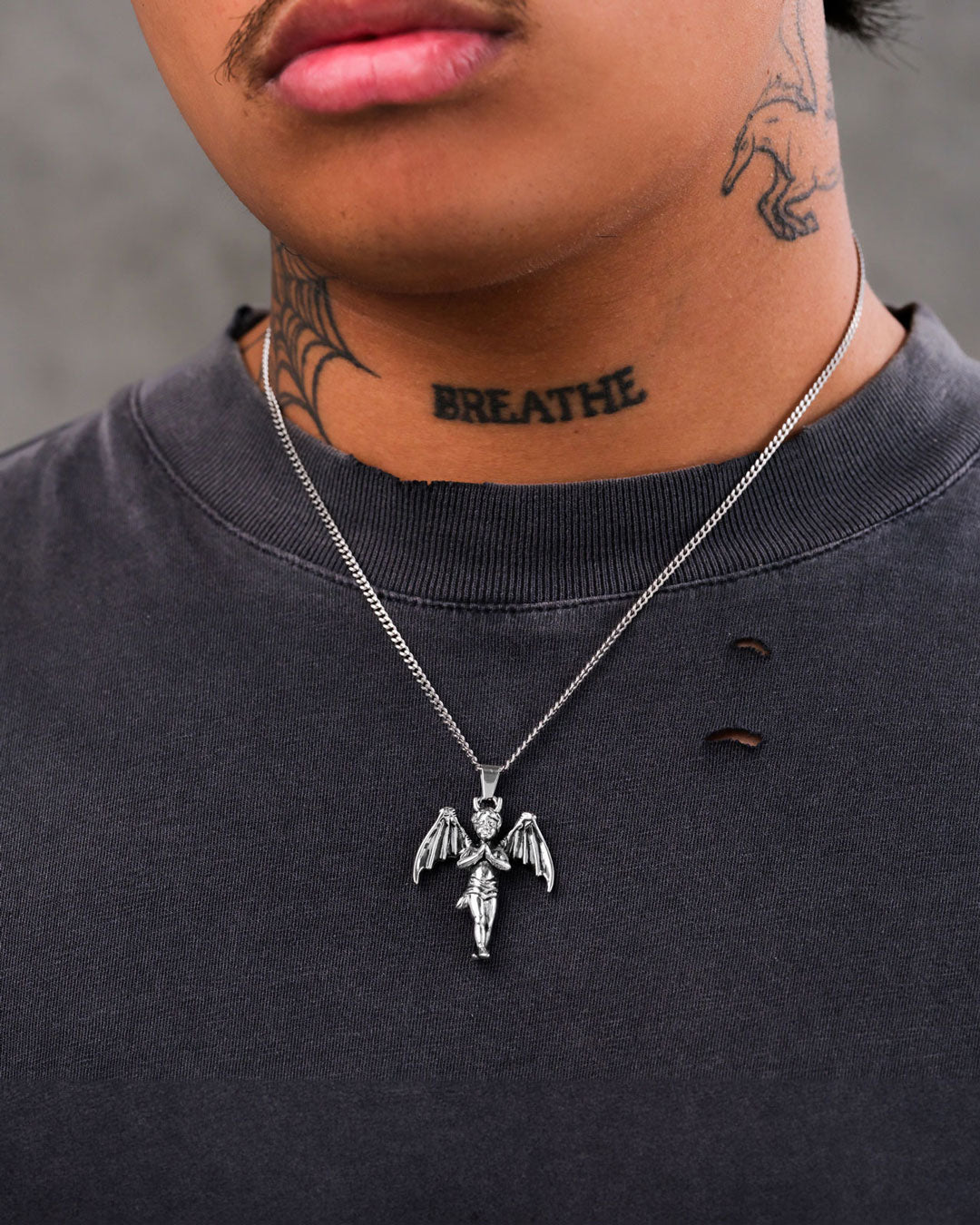 A person wearing a black shirt with small holes, "The Wicked One" Pendant in stainless steel, and neck tattoos including the word BREATHE and a design reaching up to the jawline.