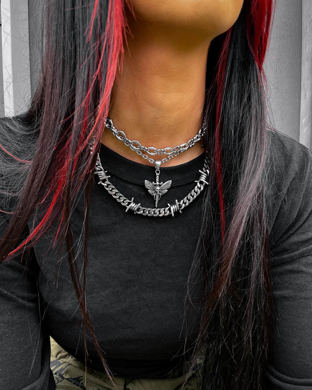 A person with long black hair and red highlights wears a black top and two silver necklaces, including the 9mm Barbed Wire Cuban Chain with a dragonfly pendant. The photo is cropped just below the nose.
